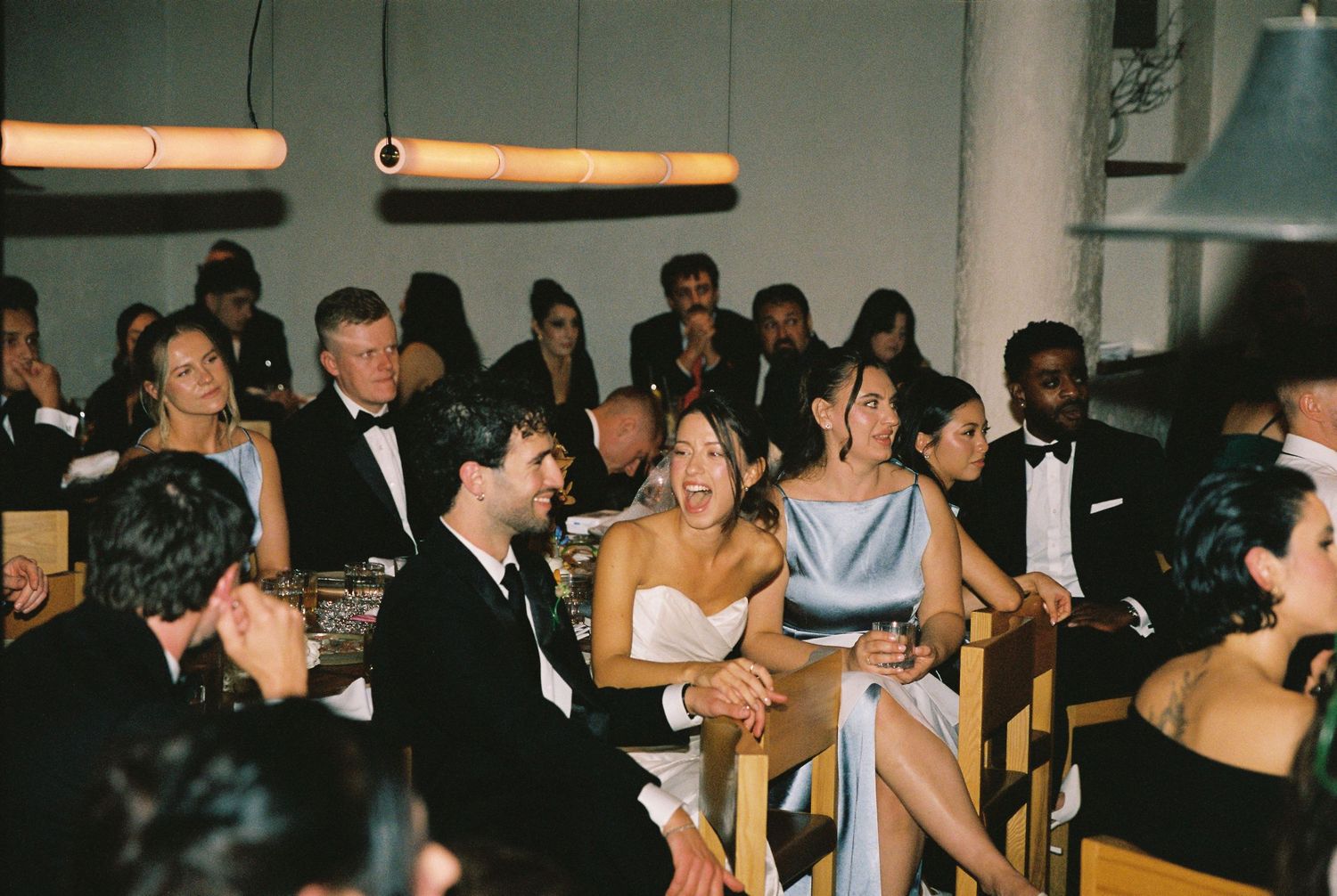 Wedding guests laugh and celebrate together at a modern indoor reception venue with warm pendant lighting.