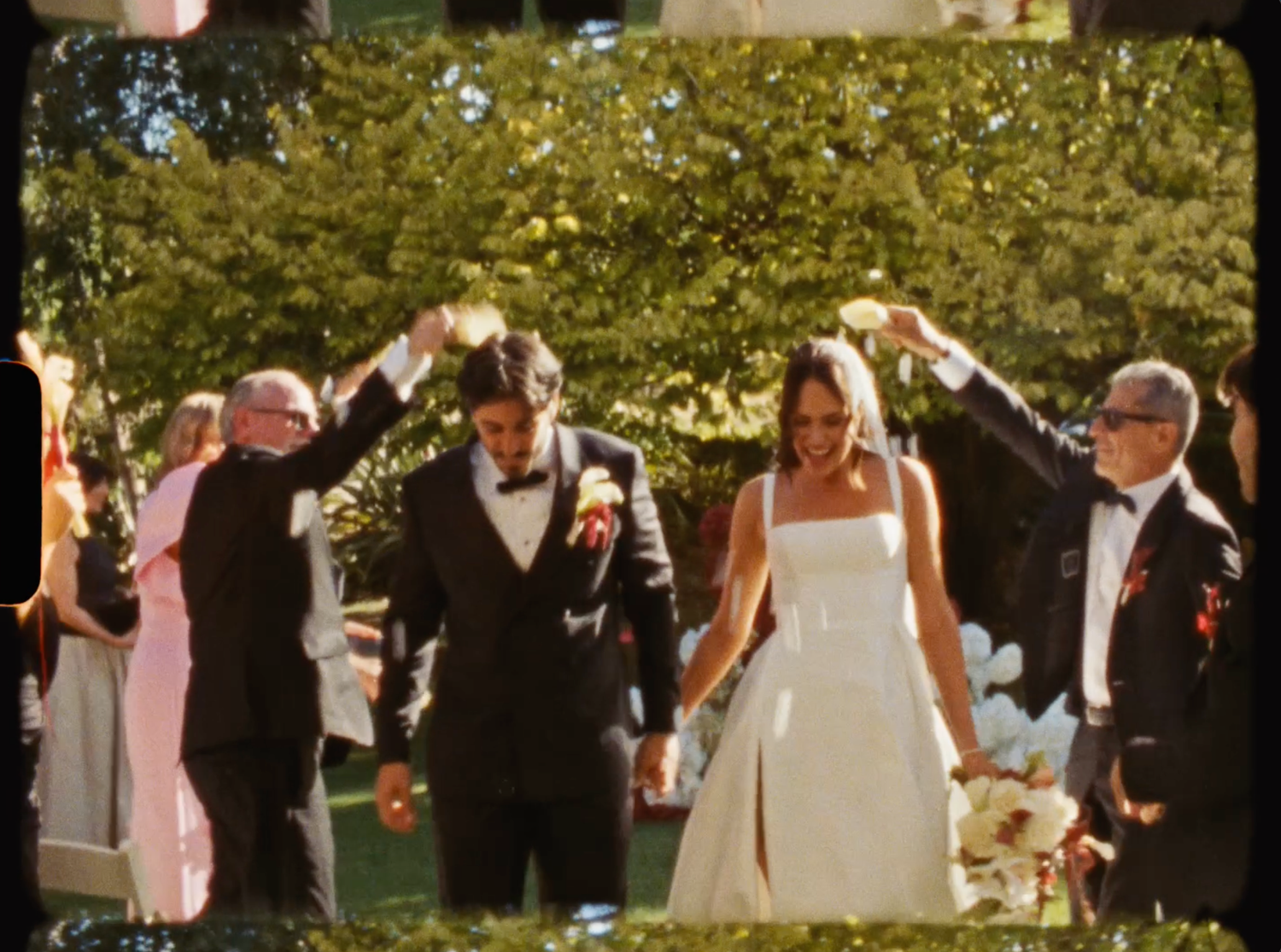 Newlyweds walking through a celebratory arch formed by guests after outdoor garden wedding ceremony.