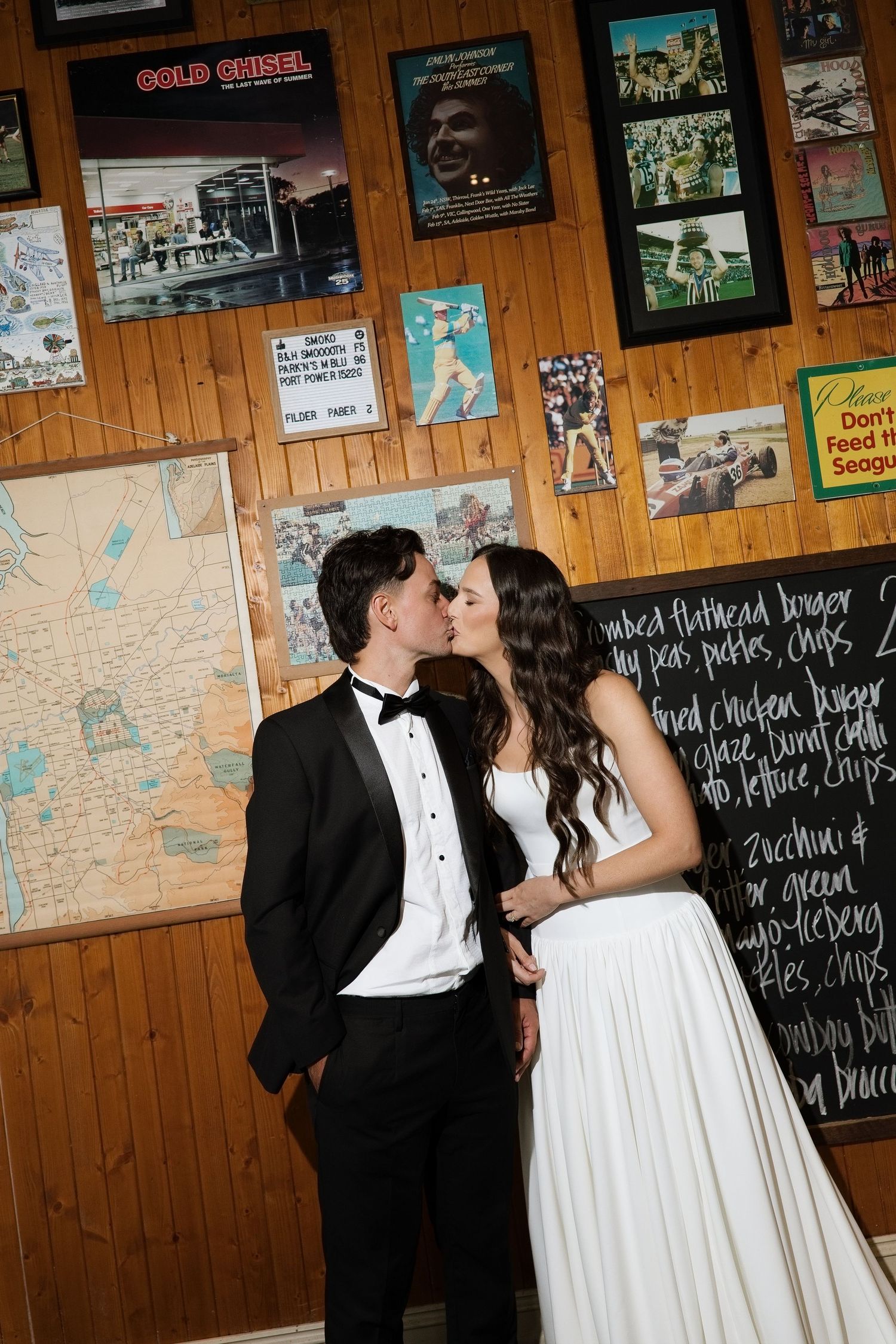 Bride and groom sharing a romantic kiss in front of a wood-paneled wall covered with vintage posters.