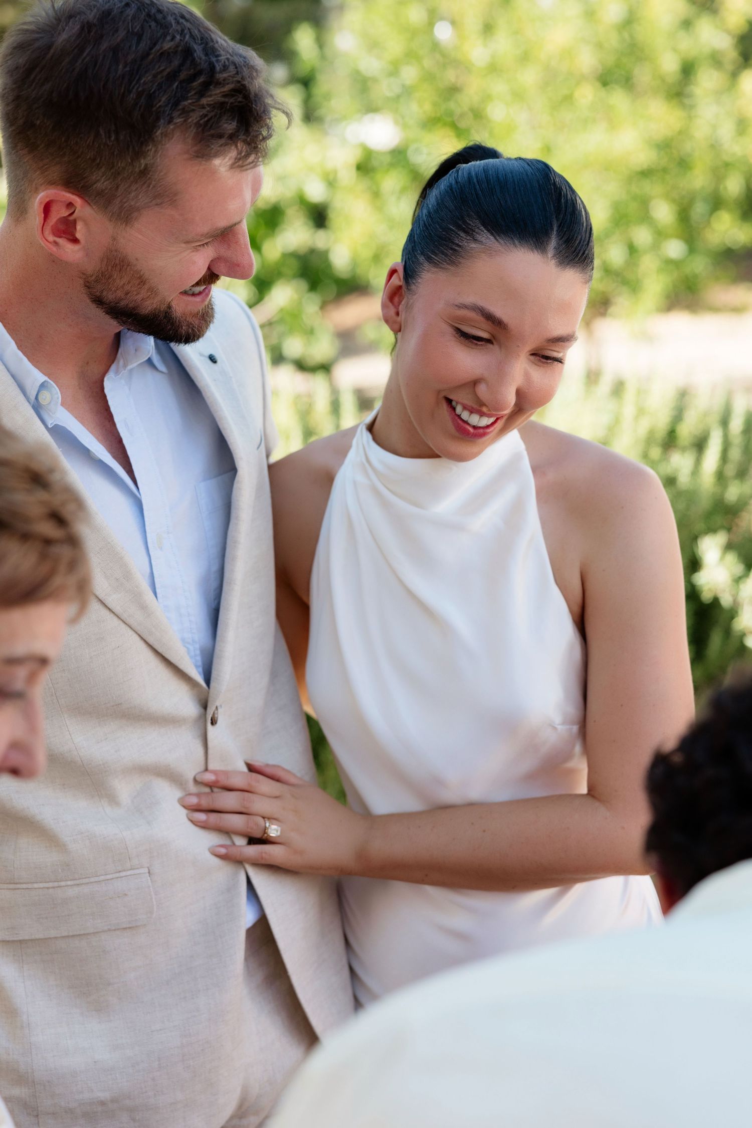 Smiling bride in white halter gown holding grooms arm during outdoor wedding reception.