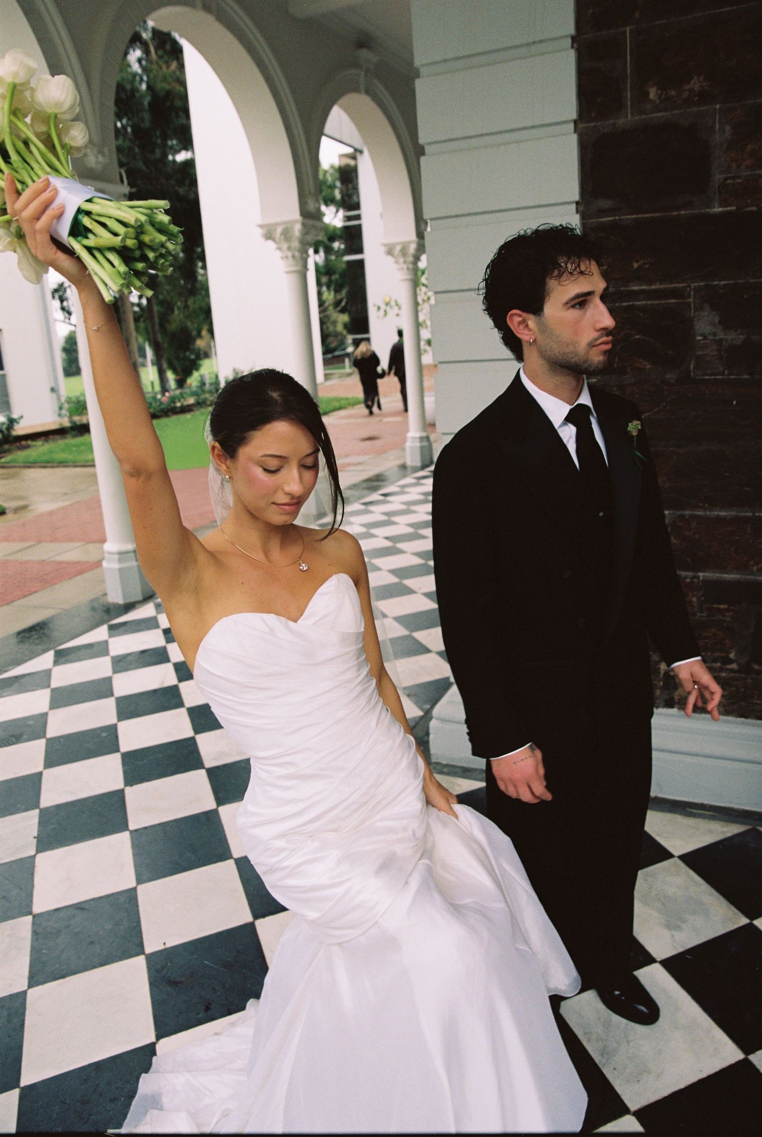 Bride in strapless white mermaid gown raises bouquet while groom stands beside her on checkered tile.