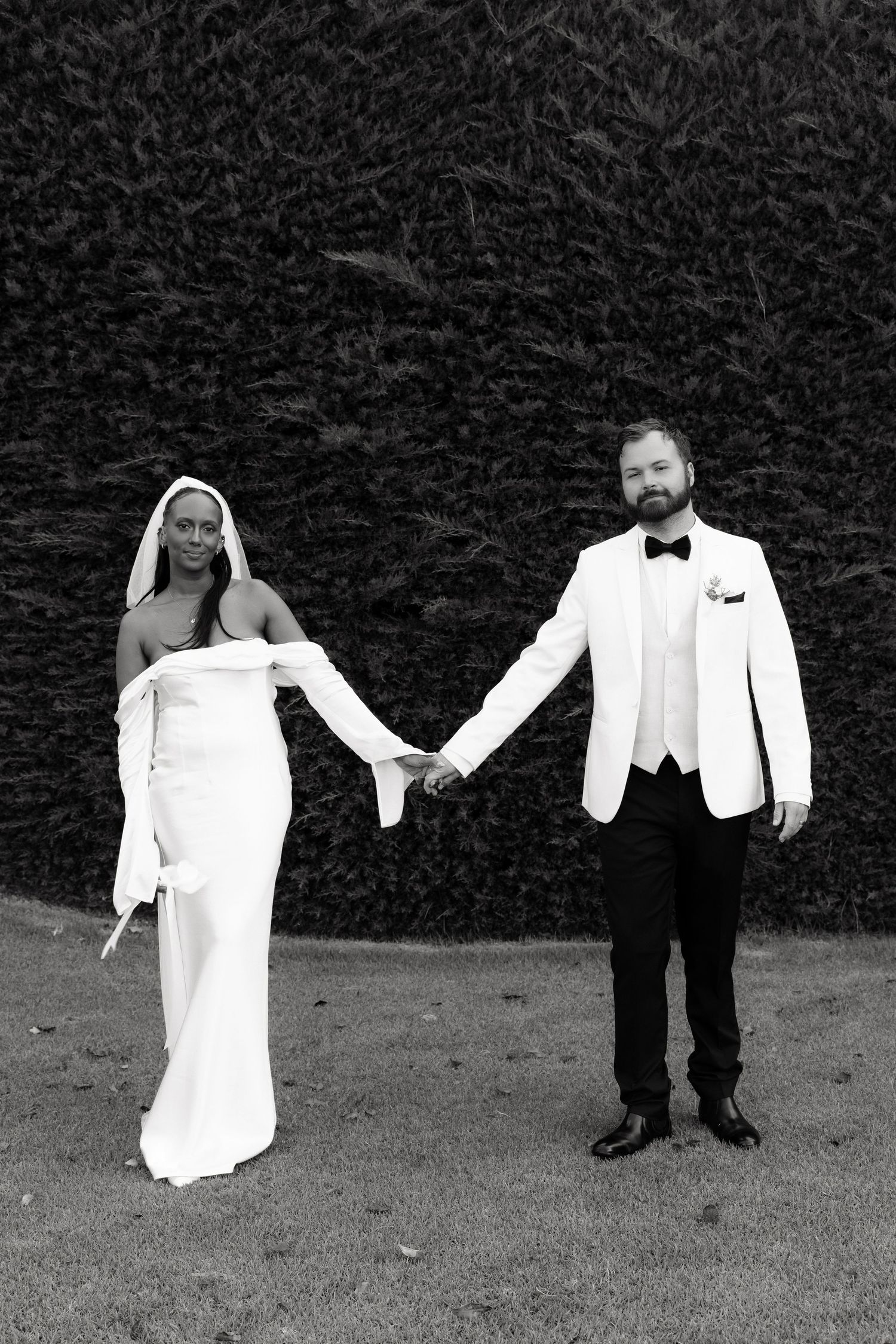 Black and white portrait of bride in strapless gown and groom in white tuxedo holding hands outdoors.