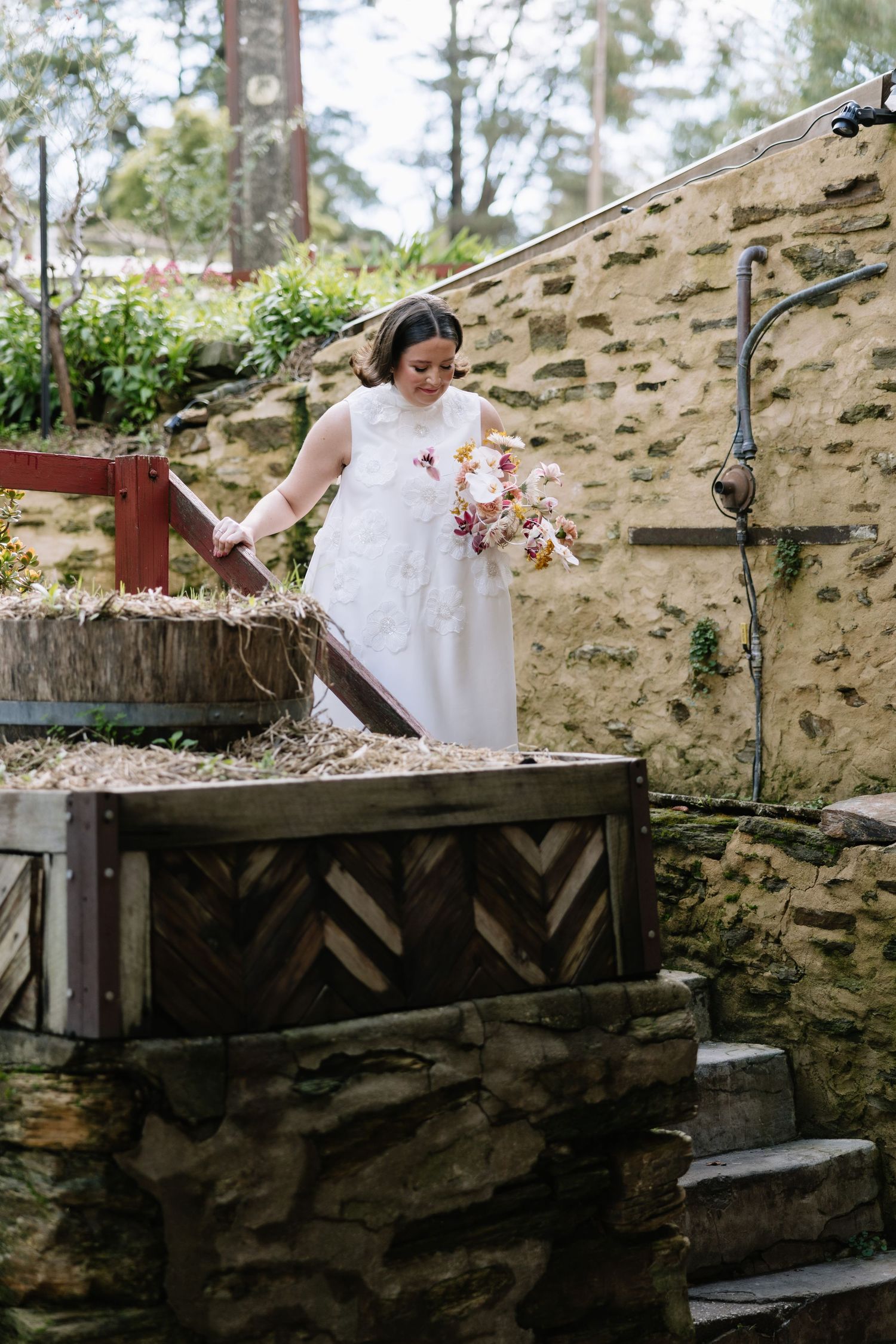 Bride in white gown holding autumn bouquet near rustic stone wall and wooden water feature outdoors.
