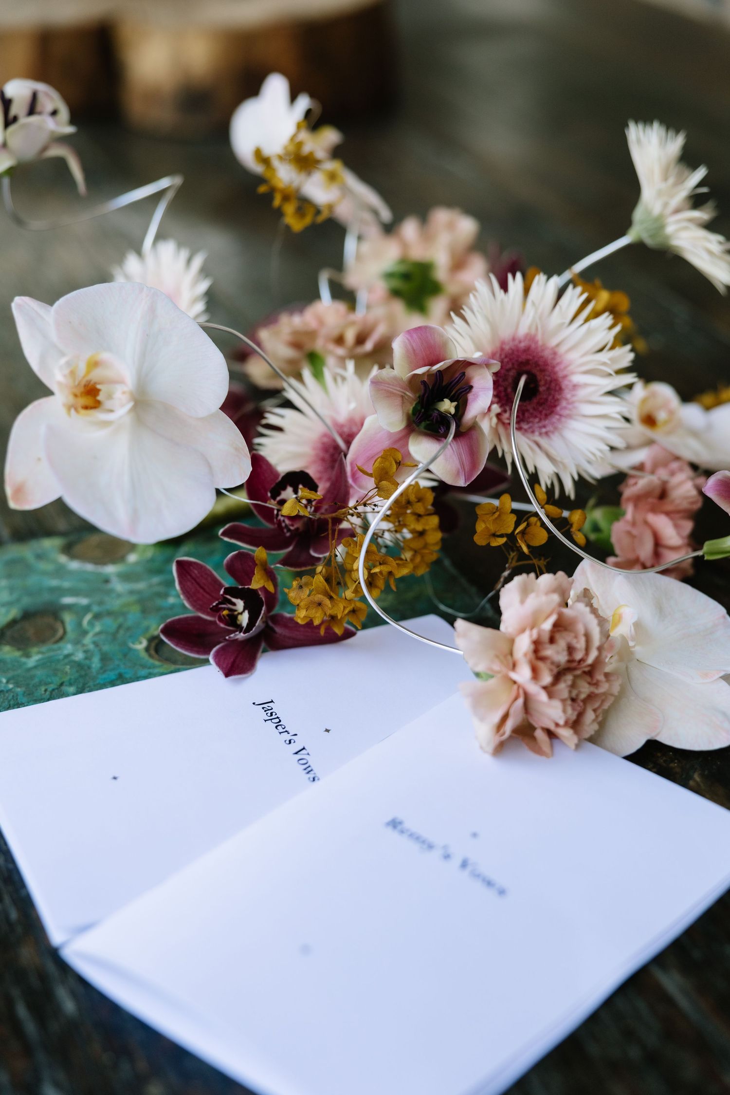 Wedding vows booklets beside a lush floral arrangement of orchids, gerberas, and carnations in blush and burgundy tones.
