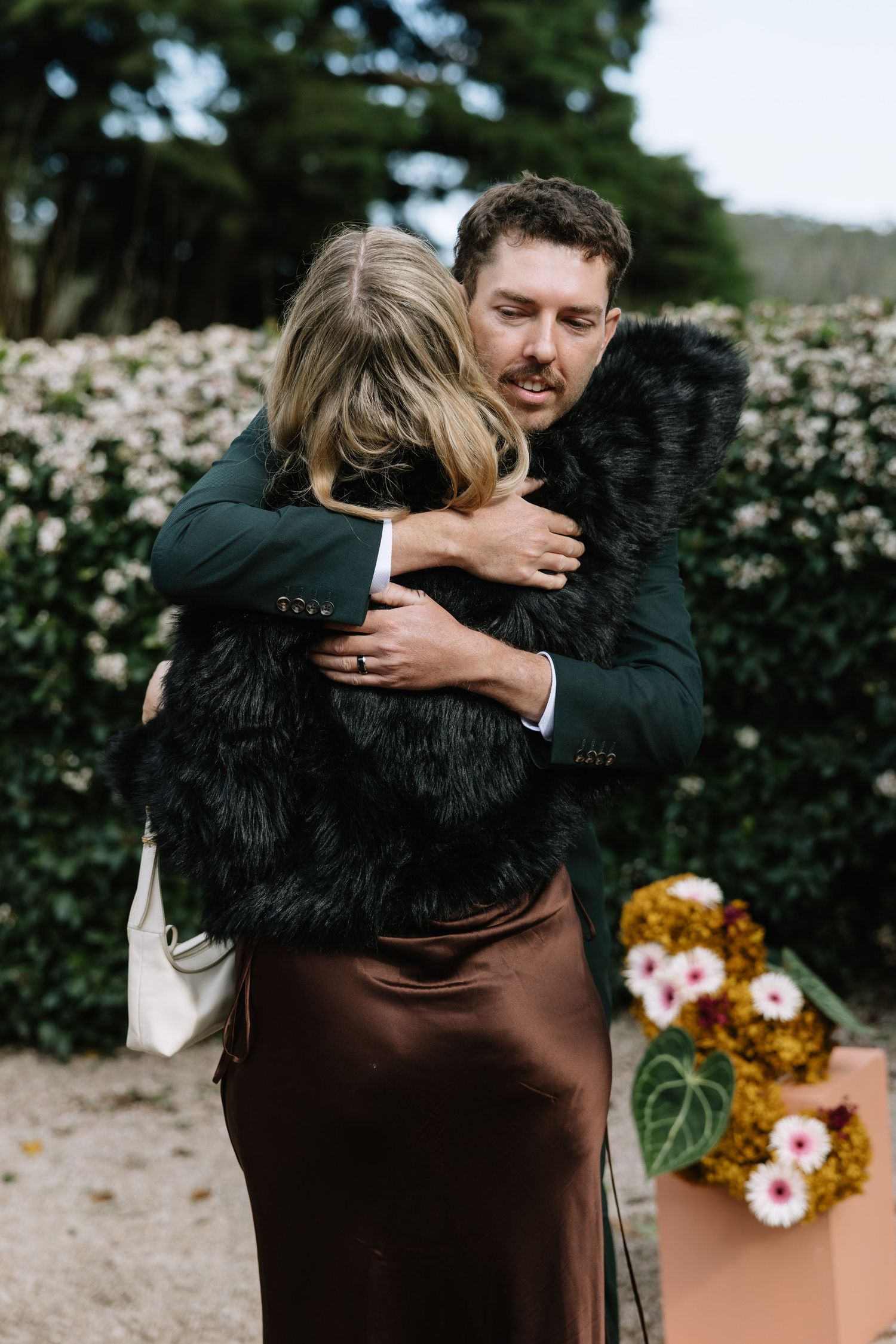 A man in a dark suit hugs a woman in a black fur vest and brown satin skirt outdoors near flowering hedges.