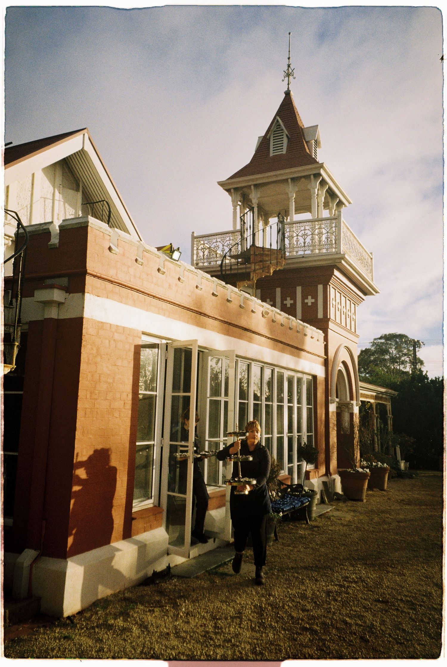 A waiter serves guests outside a historic Victorian brick building with an ornate tower and large windows on a sunny day.