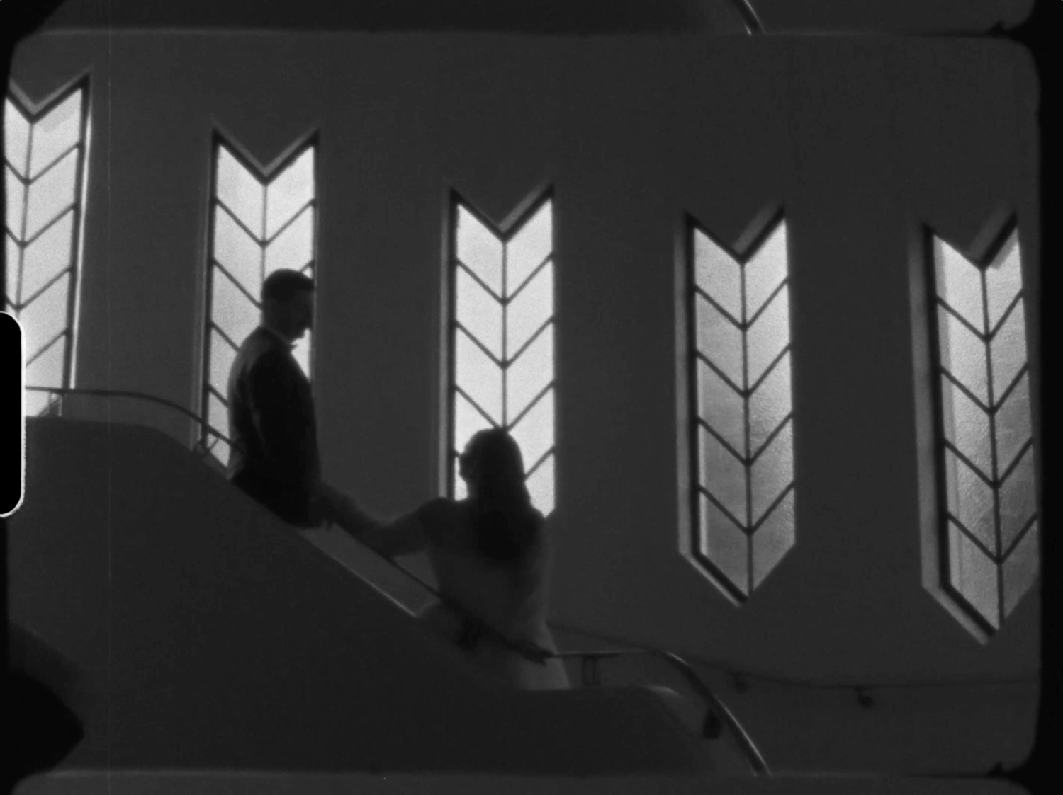 Black and white silhouette of two people on stairs beside decorative arrow-shaped stained glass windows.
