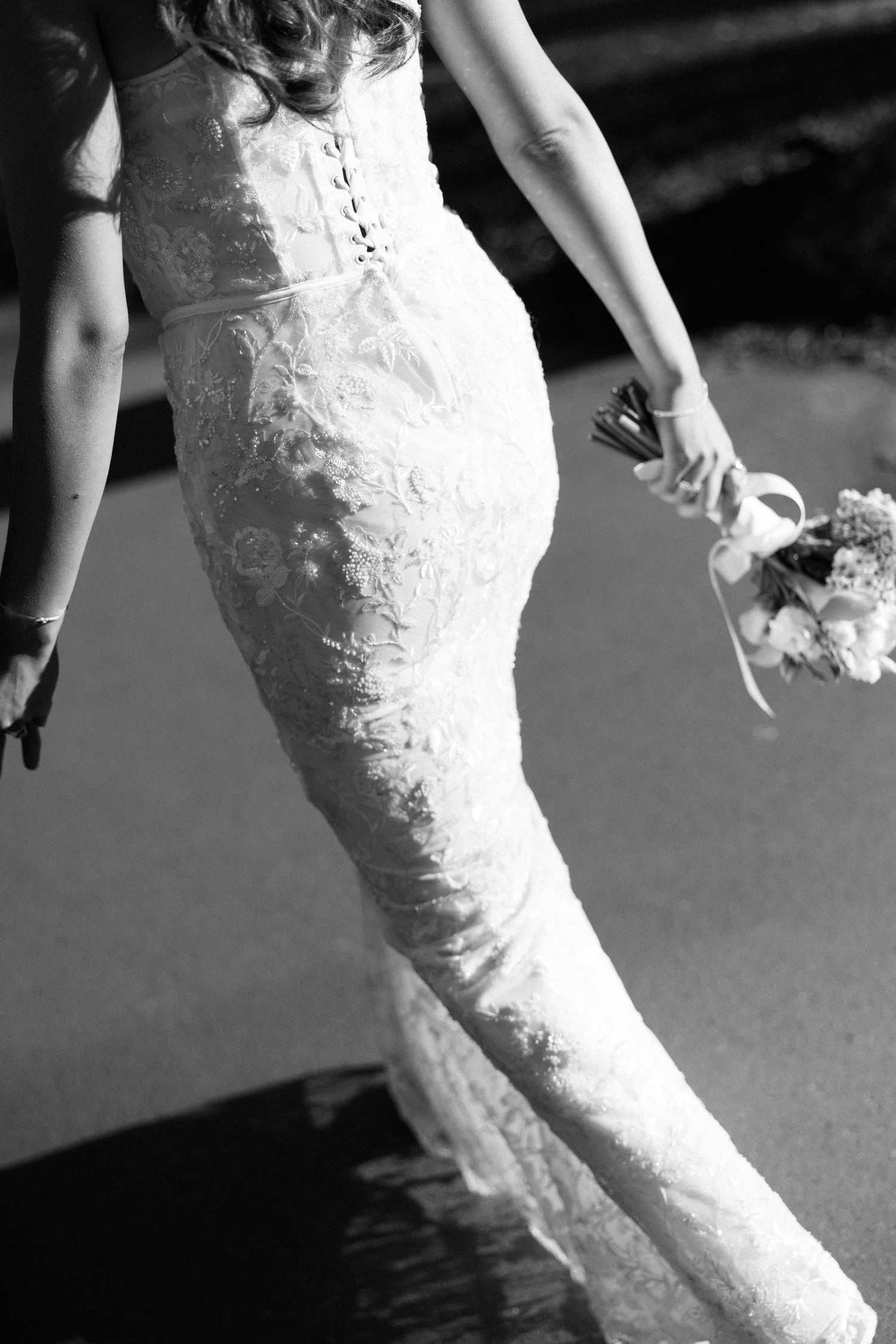 Black and white photo of a bride in a lace mermaid wedding gown holding a bouquet while walking outdoors.