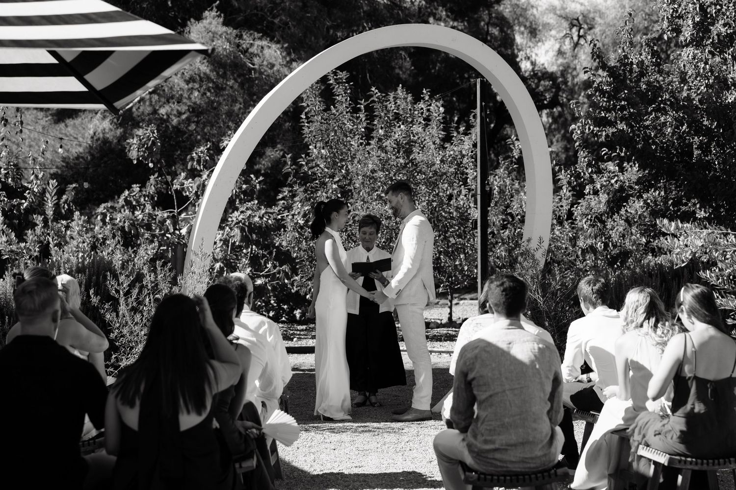 Black and white outdoor wedding ceremony under white arch with guests seated, surrounded by lush greenery.