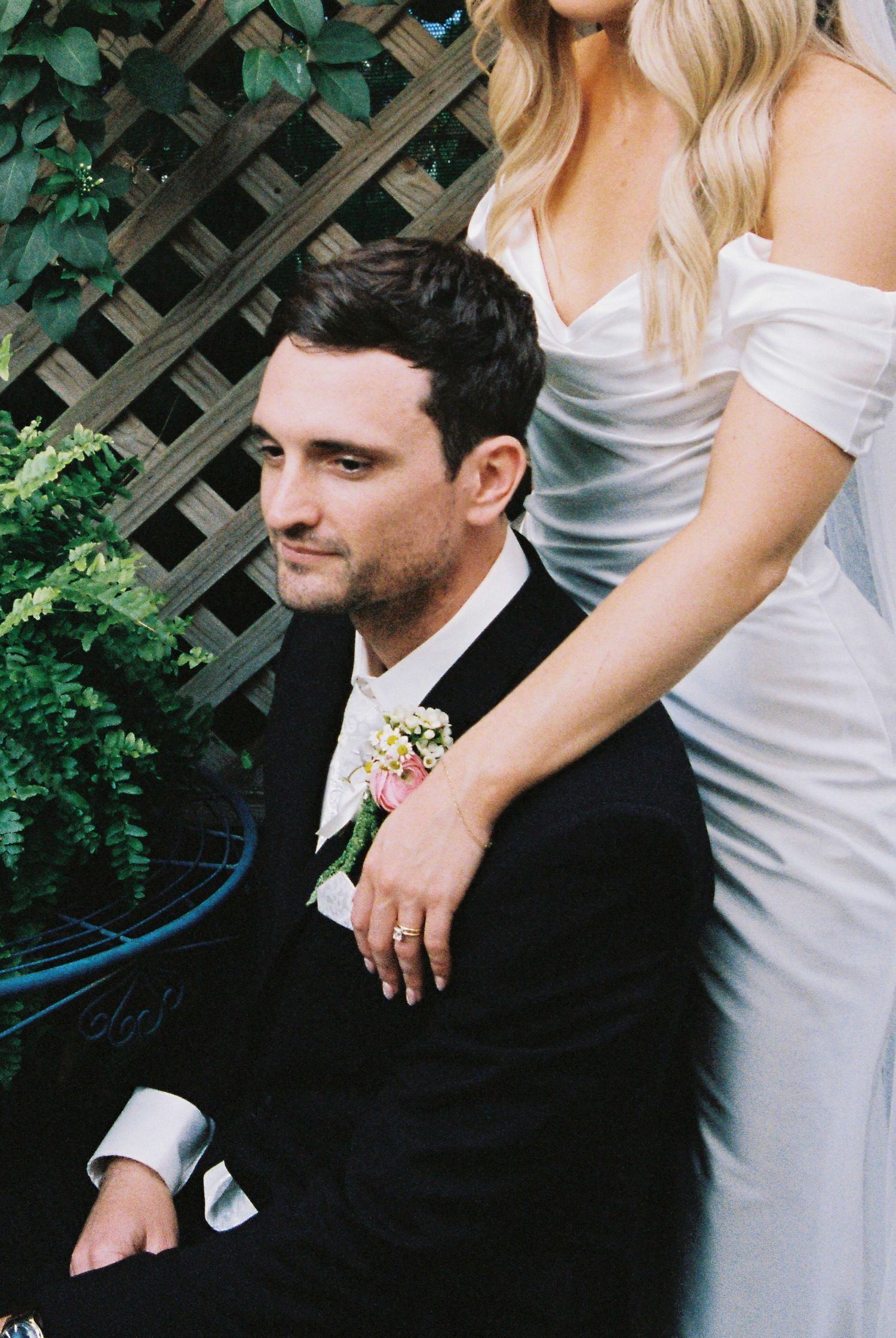 Groom seated with bride standing behind him, her hand on his shoulder, in an intimate garden portrait.