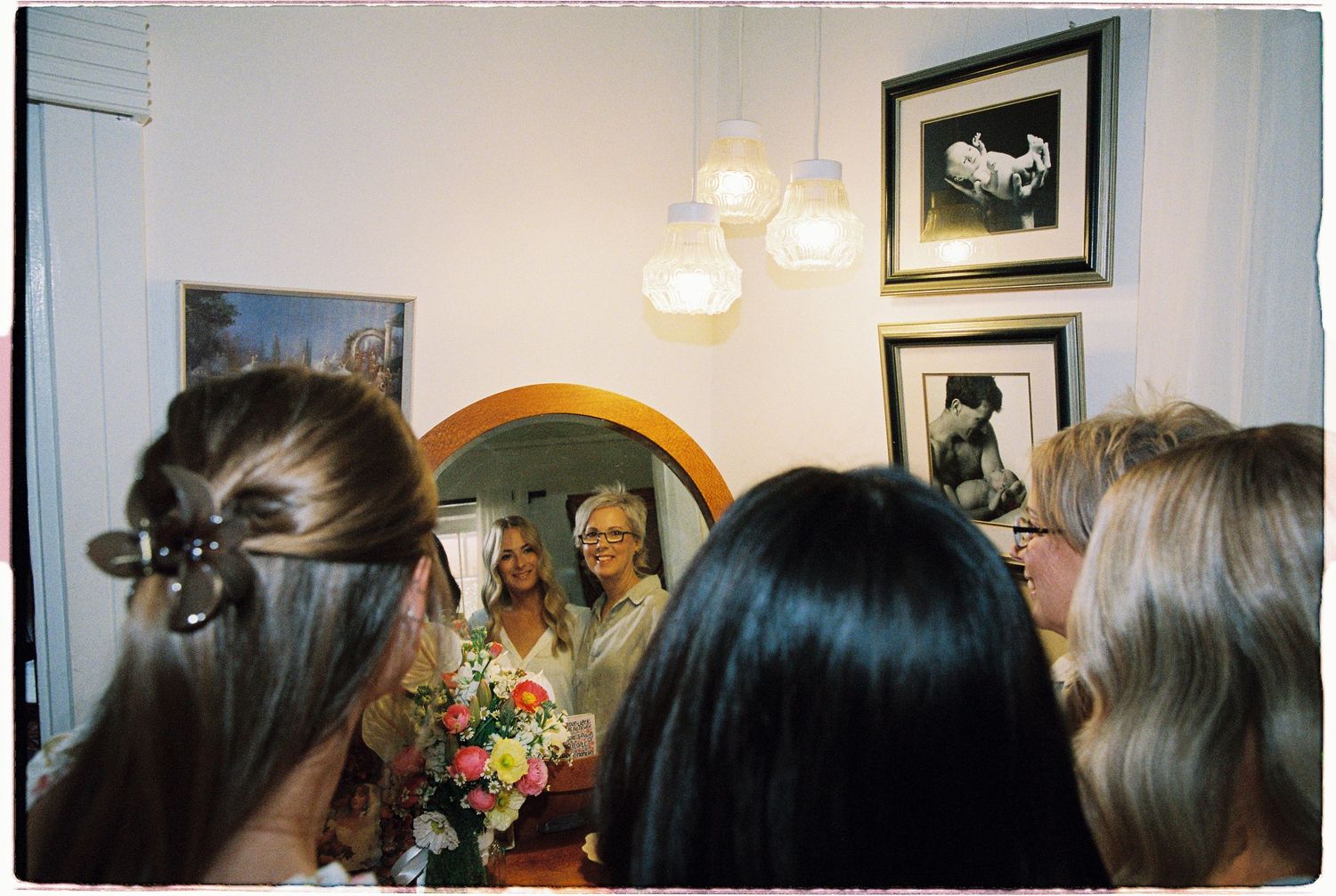 Bride holding bouquet reflected in round mirror surrounded by bridesmaids in warmly lit room with framed art.