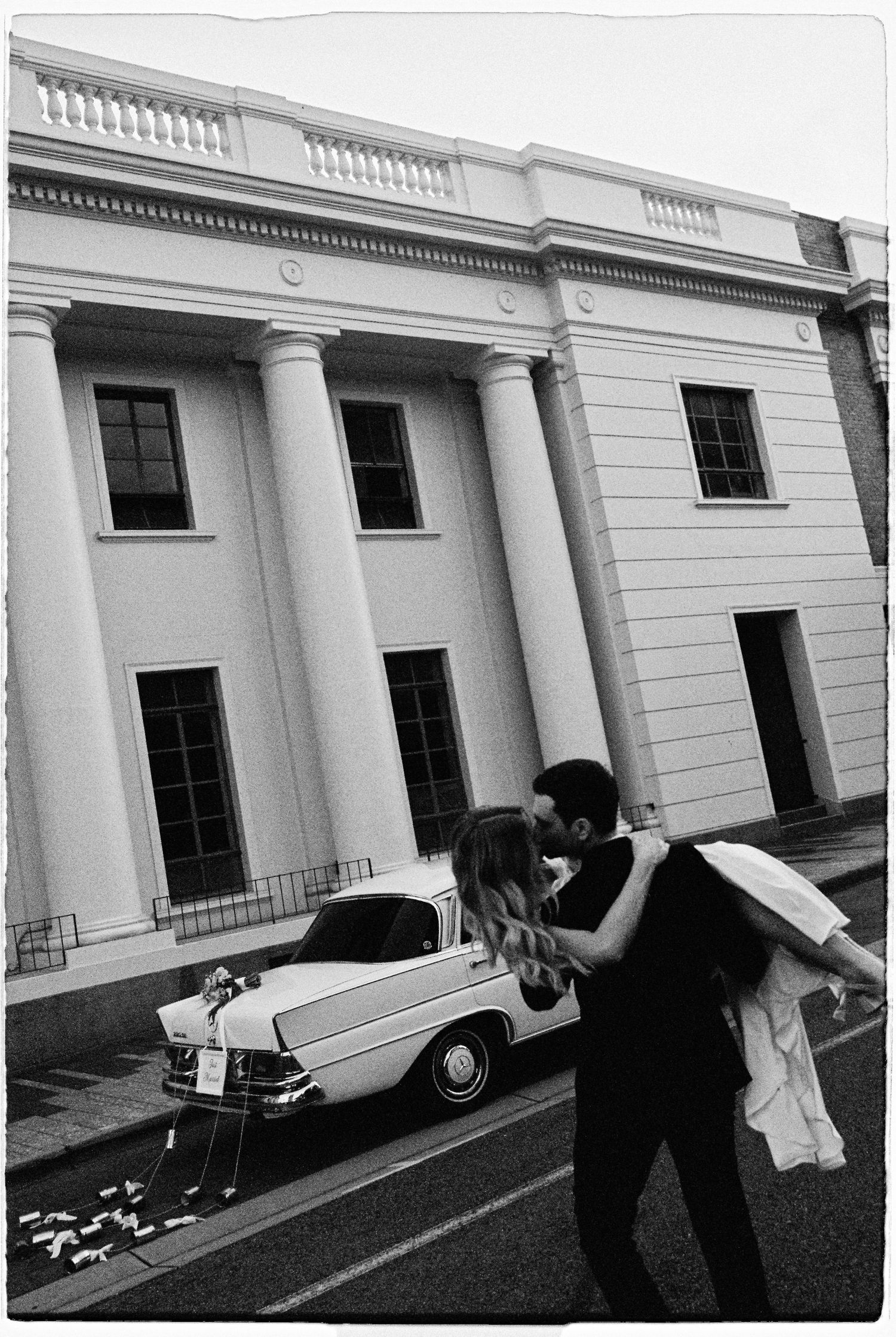 Bride and groom sharing a romantic kiss beside a vintage car in front of a grand neoclassical building.