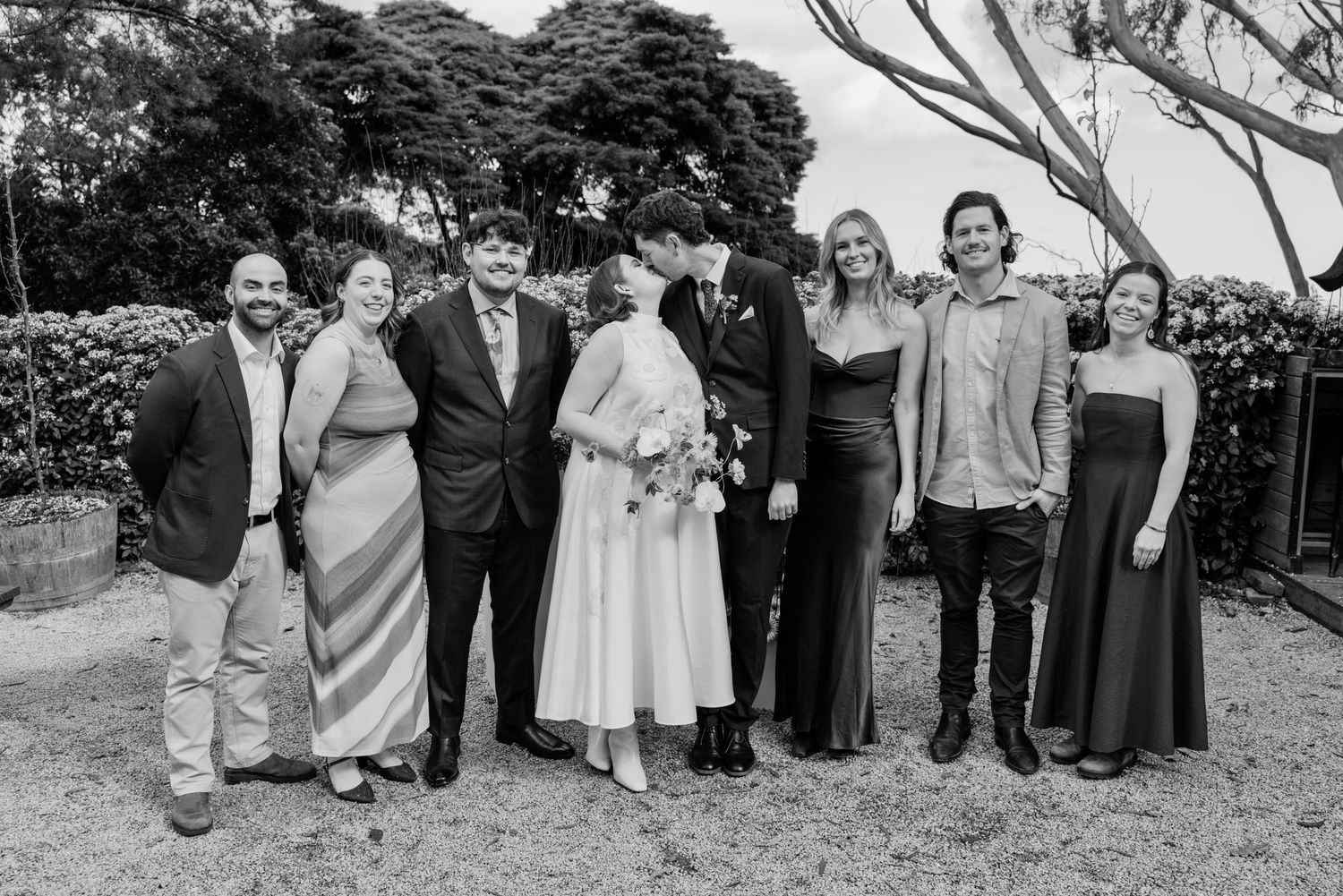 Black and white wedding photo of a bride, groom, and six guests smiling together outdoors at a garden venue.
