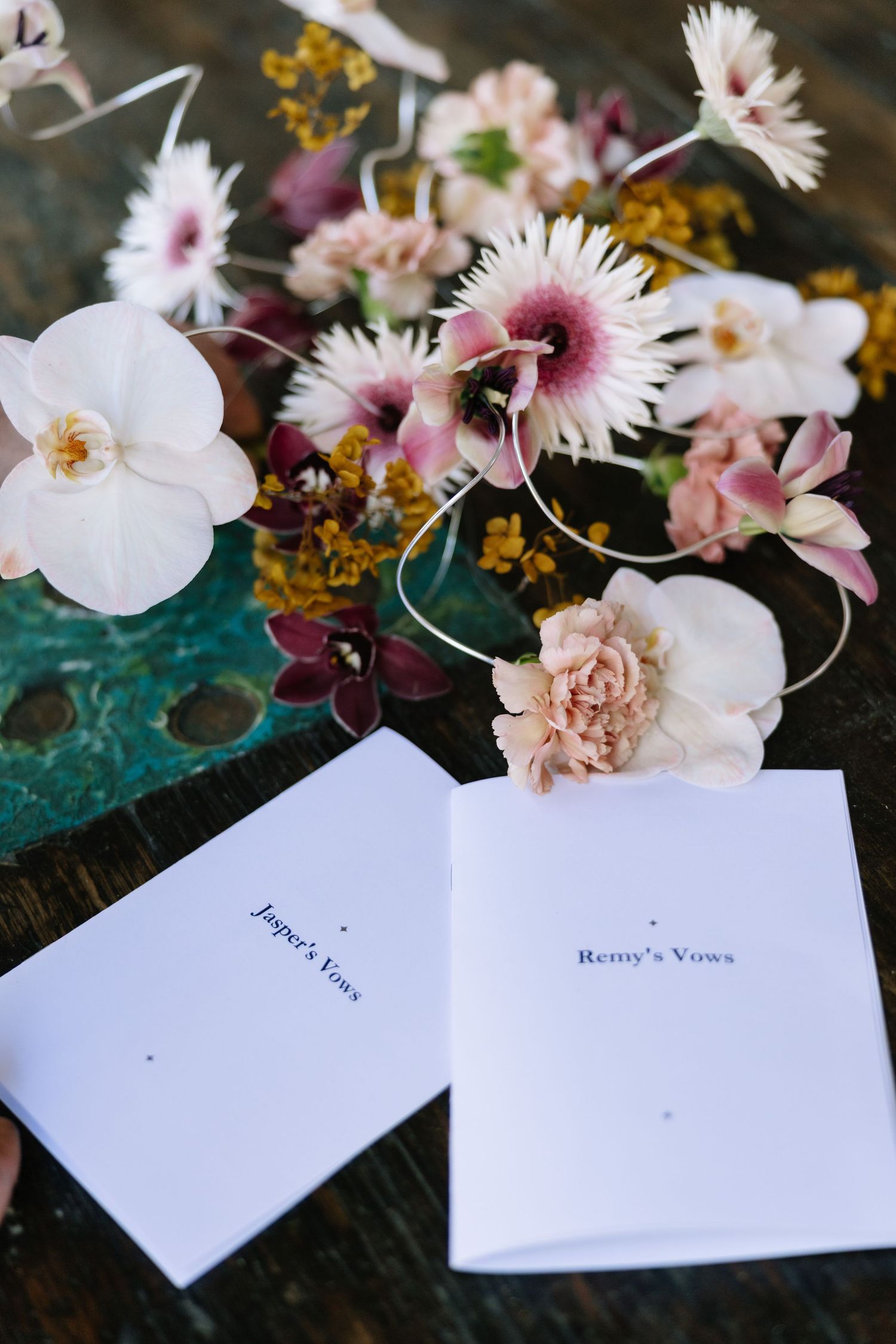 Wedding vow booklets for Jasper and Remy surrounded by pink, white, and yellow flowers on a rustic wooden table.