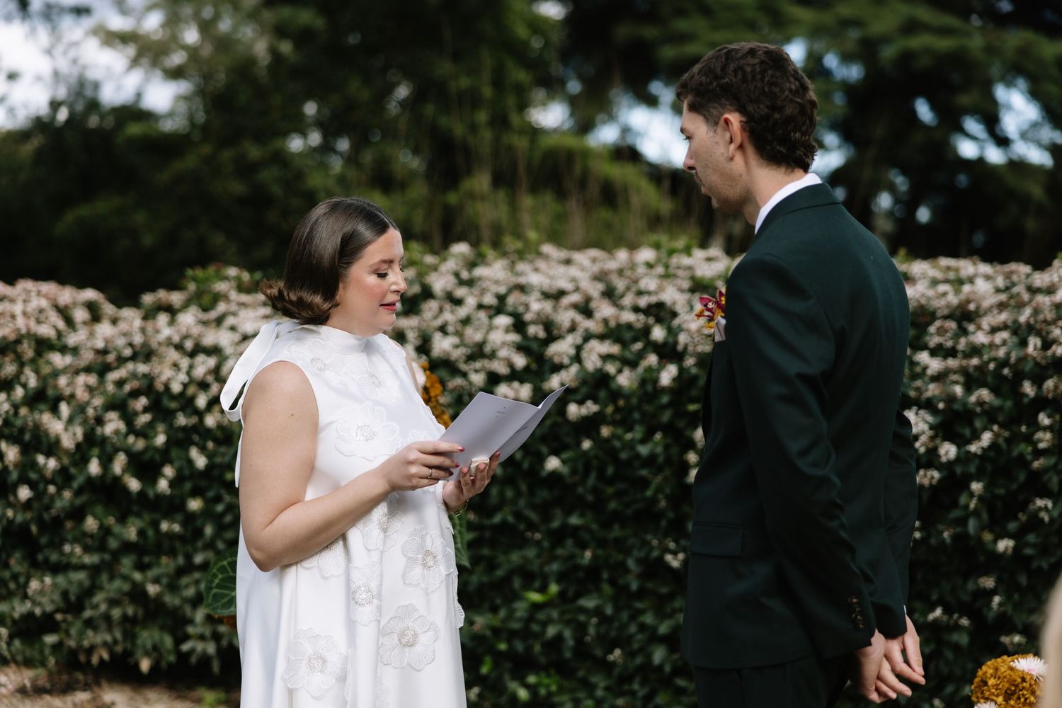 Bride reads wedding vows to groom outdoors, surrounded by lush white flowering hedges at an elegant garden ceremony.