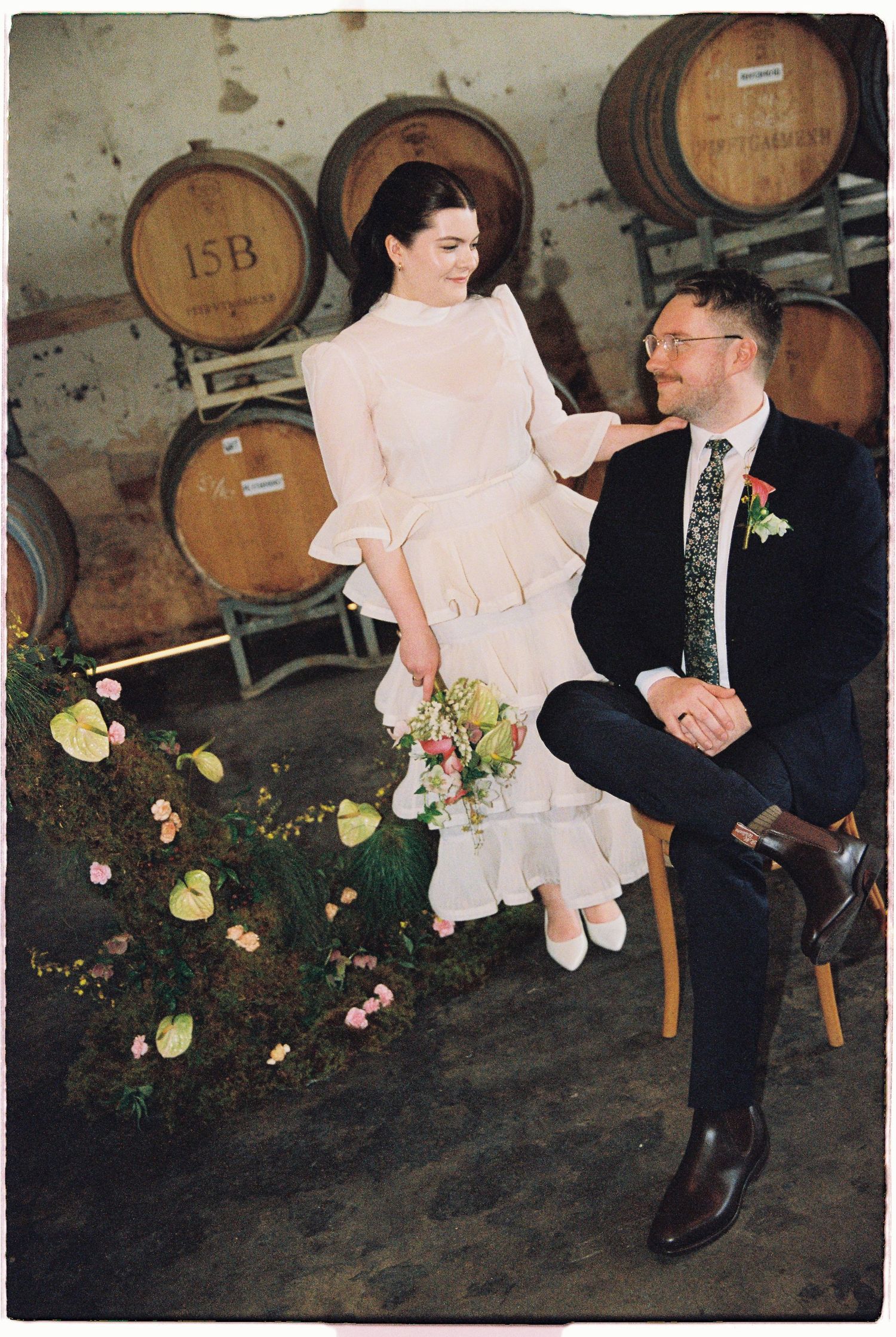 Bride and groom sit together among wine barrels in a winery ceremony space, bride holding a colorful bouquet.