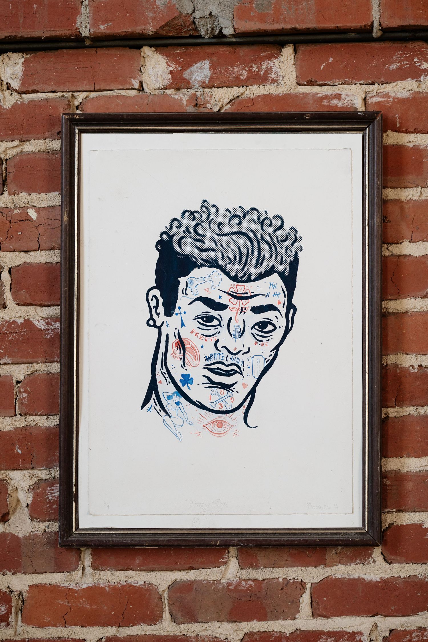 Framed illustration of a tattooed man with curly hair hanging on a rustic red brick wall.