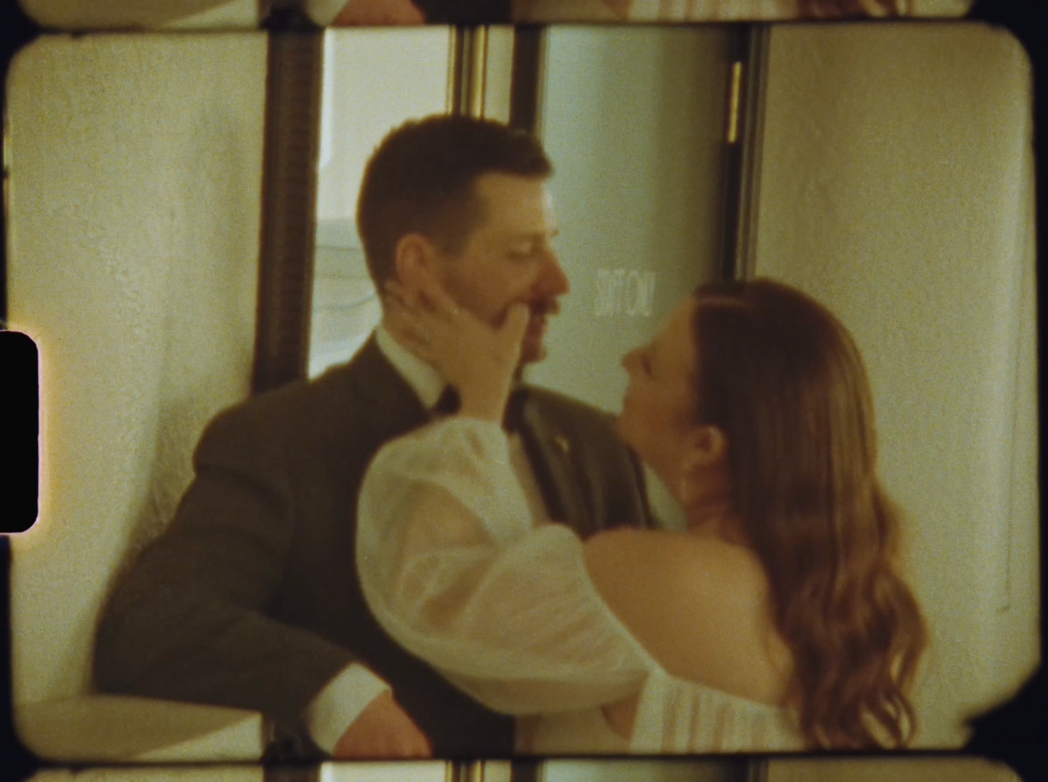A couple shares a romantic moment, smiling and embracing near a mirror in soft vintage film photography style.