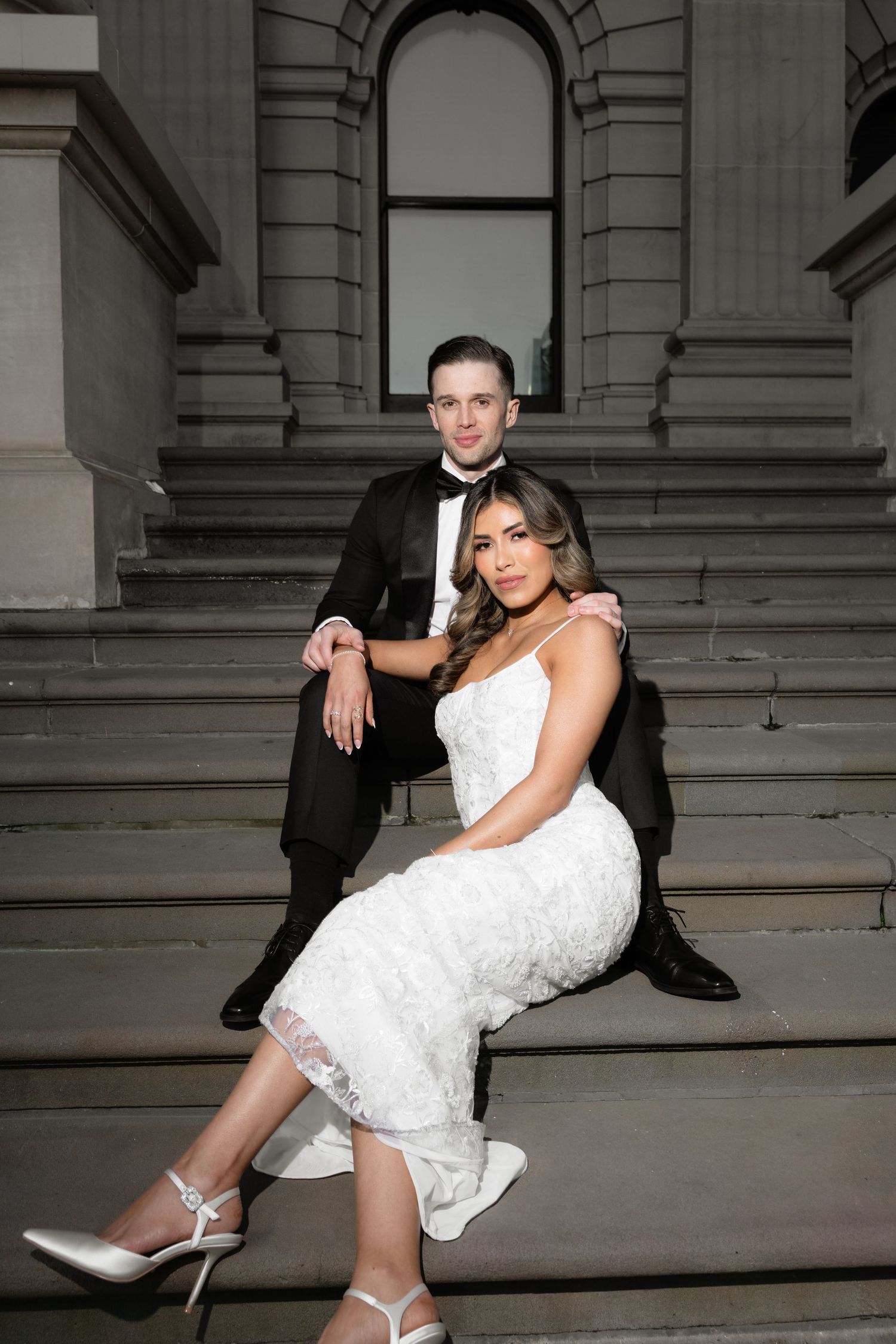 Bride and groom posing on stone steps of grand building, she in white lace dress, he in black tuxedo.