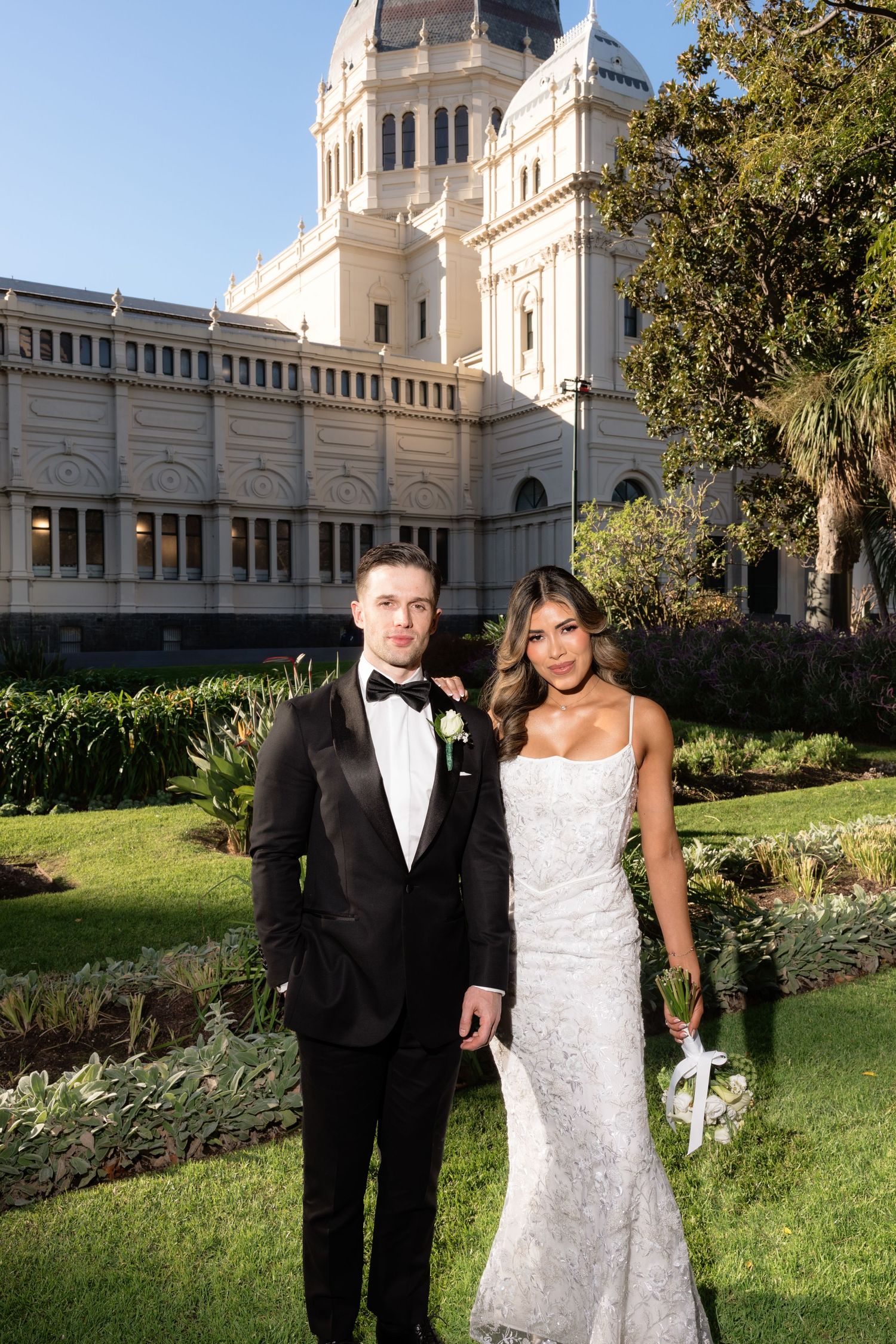 Bride and groom posing in front of a grand historic building with lush gardens on their wedding day.
