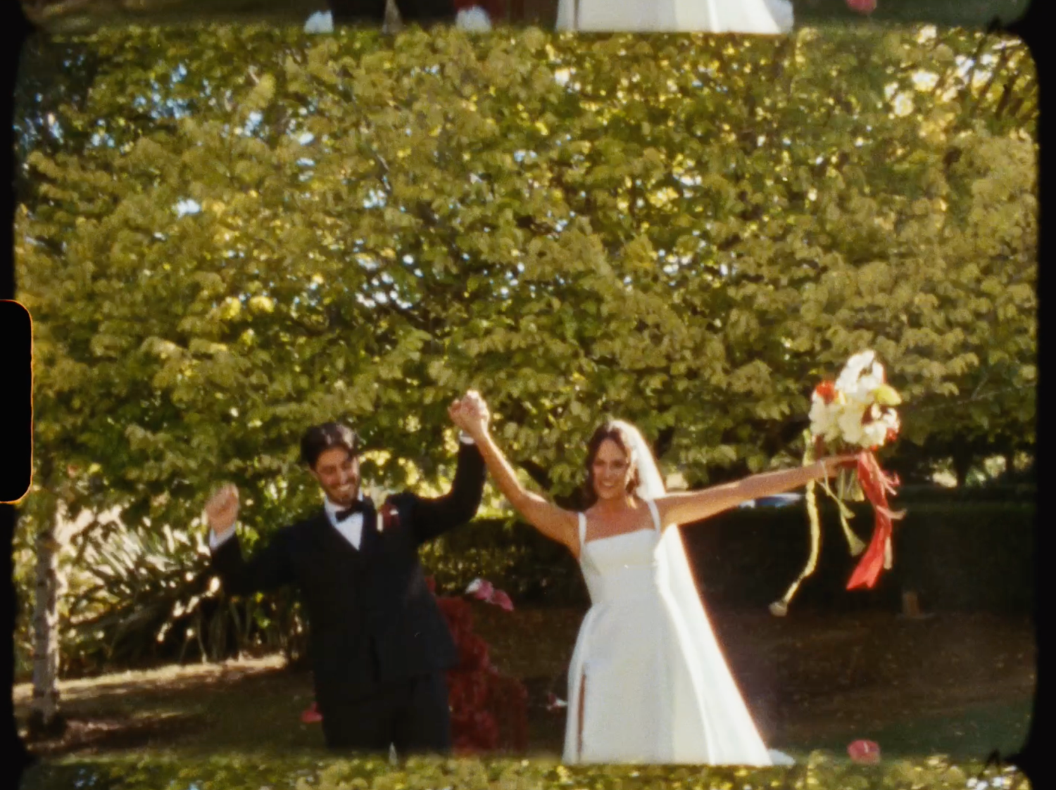 Joyful bride and groom raising hands in celebration outdoors after wedding ceremony in garden.