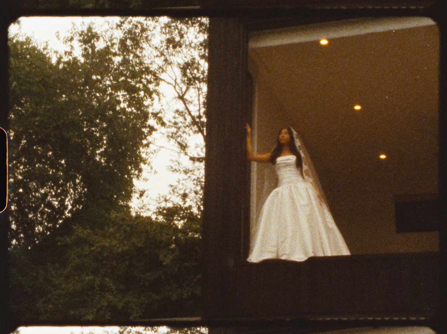 Bride in white wedding gown standing in a modern window frame surrounded by lush green trees.