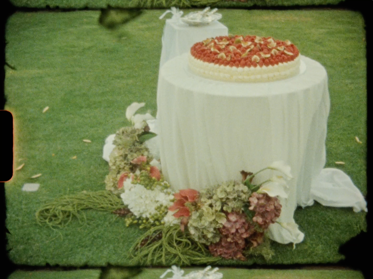 Wedding cake decorated with strawberries on a white draped table surrounded by floral arrangements on green grass.