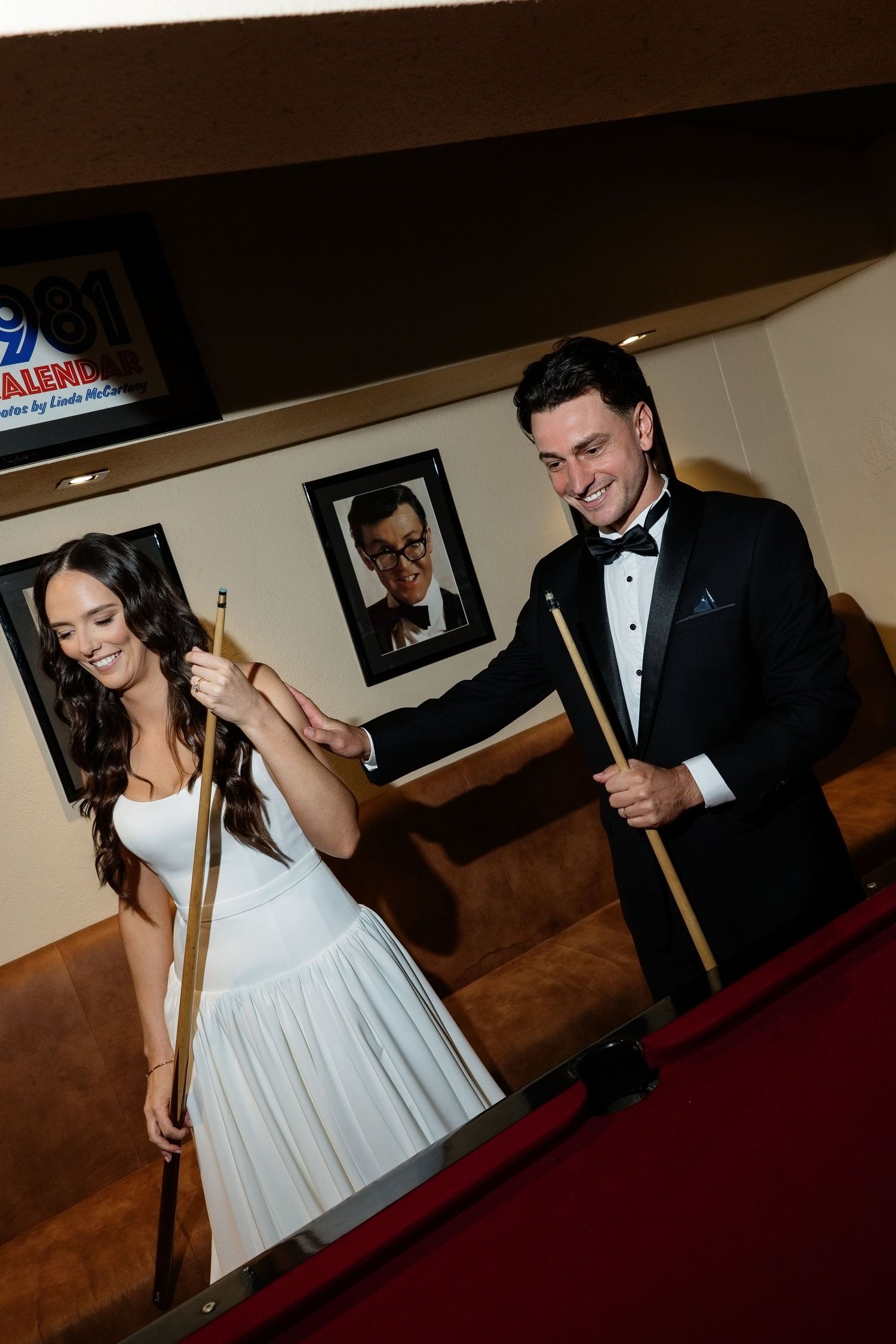 Smiling bride and groom in formal wedding attire holding pool cues together beside a billiard table.