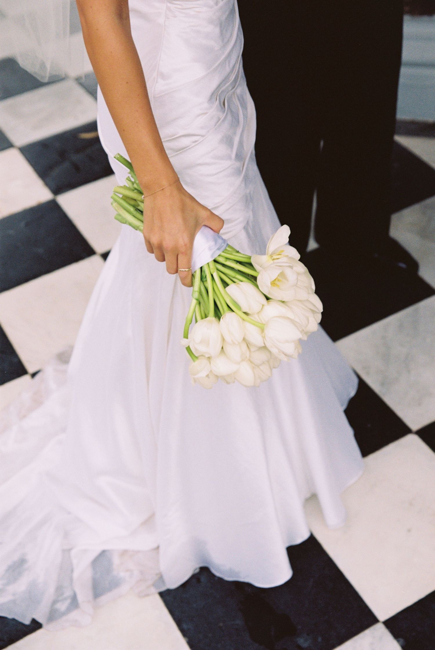 Close-up of bride in white wedding dress holding elegant white tulip bouquet on checkered floor.
