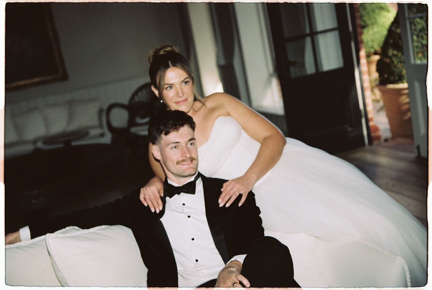 Bride in strapless white gown posing with groom in tuxedo on elegant sofa.