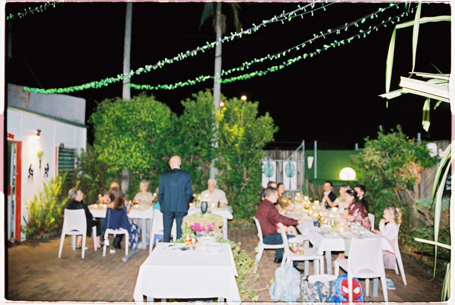 Outdoor evening dinner party with guests seated at white tables under string lights surrounded by tropical greenery.