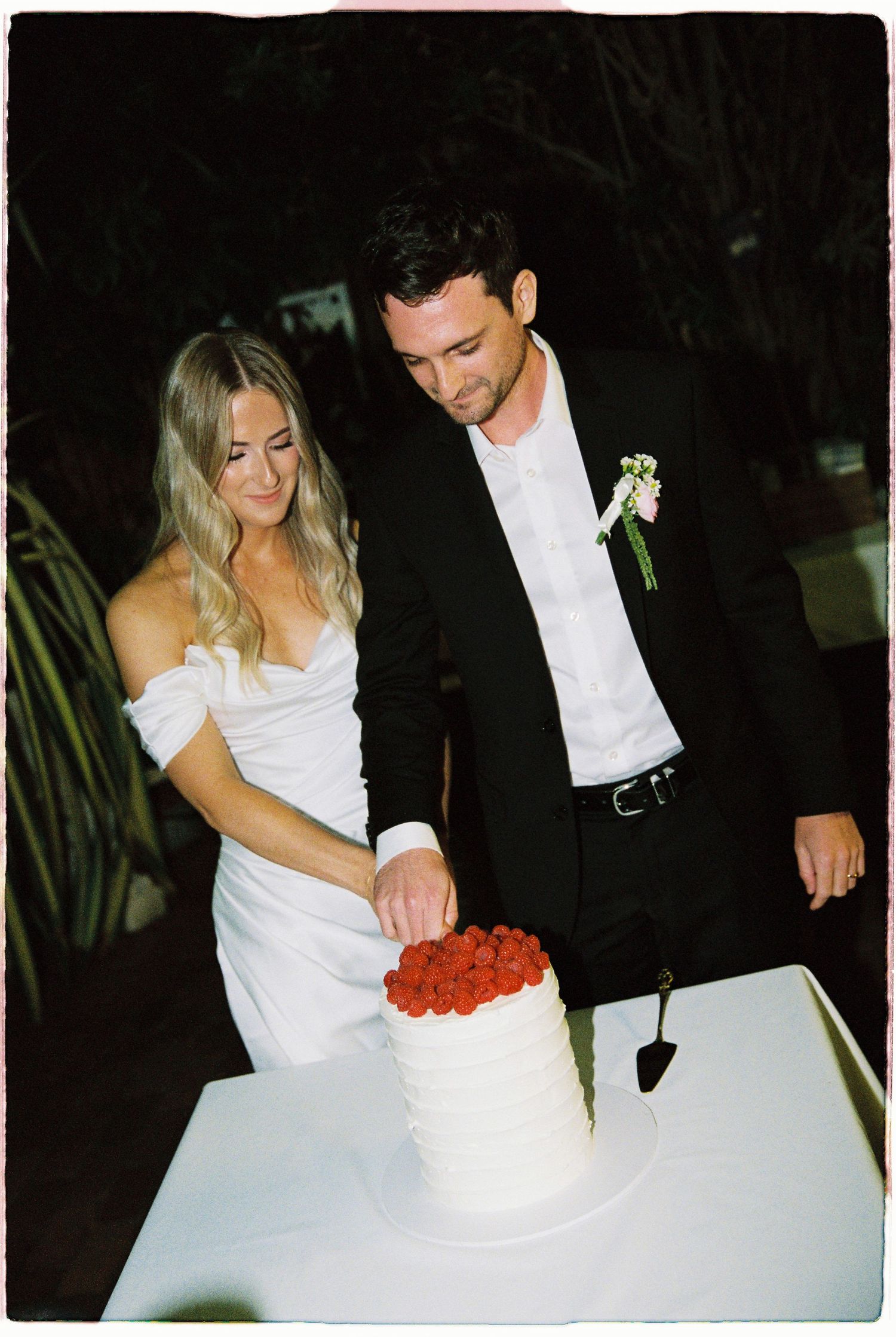 Bride and groom cutting white wedding cake topped with raspberries at outdoor evening wedding reception.