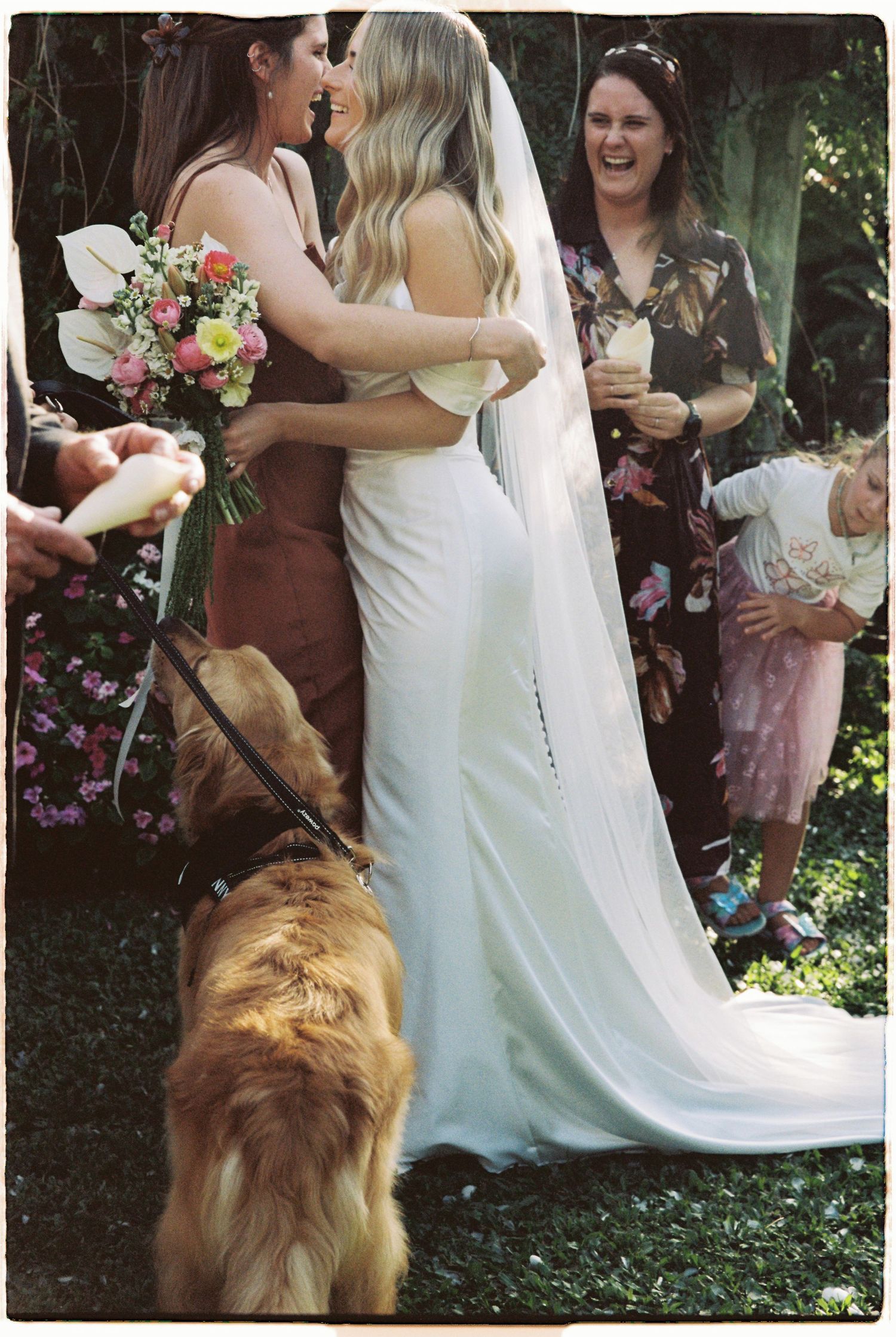 Bride in white veil hugs bridesmaid while golden retriever stands nearby.