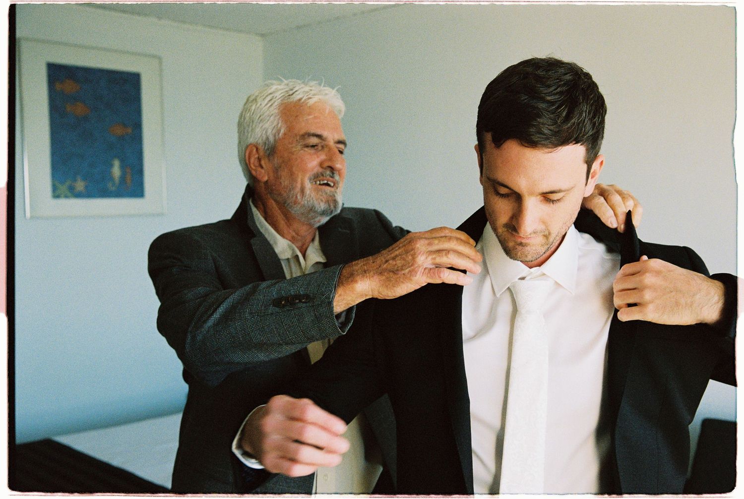 An older man helps a younger man in a suit adjust his collar indoors.