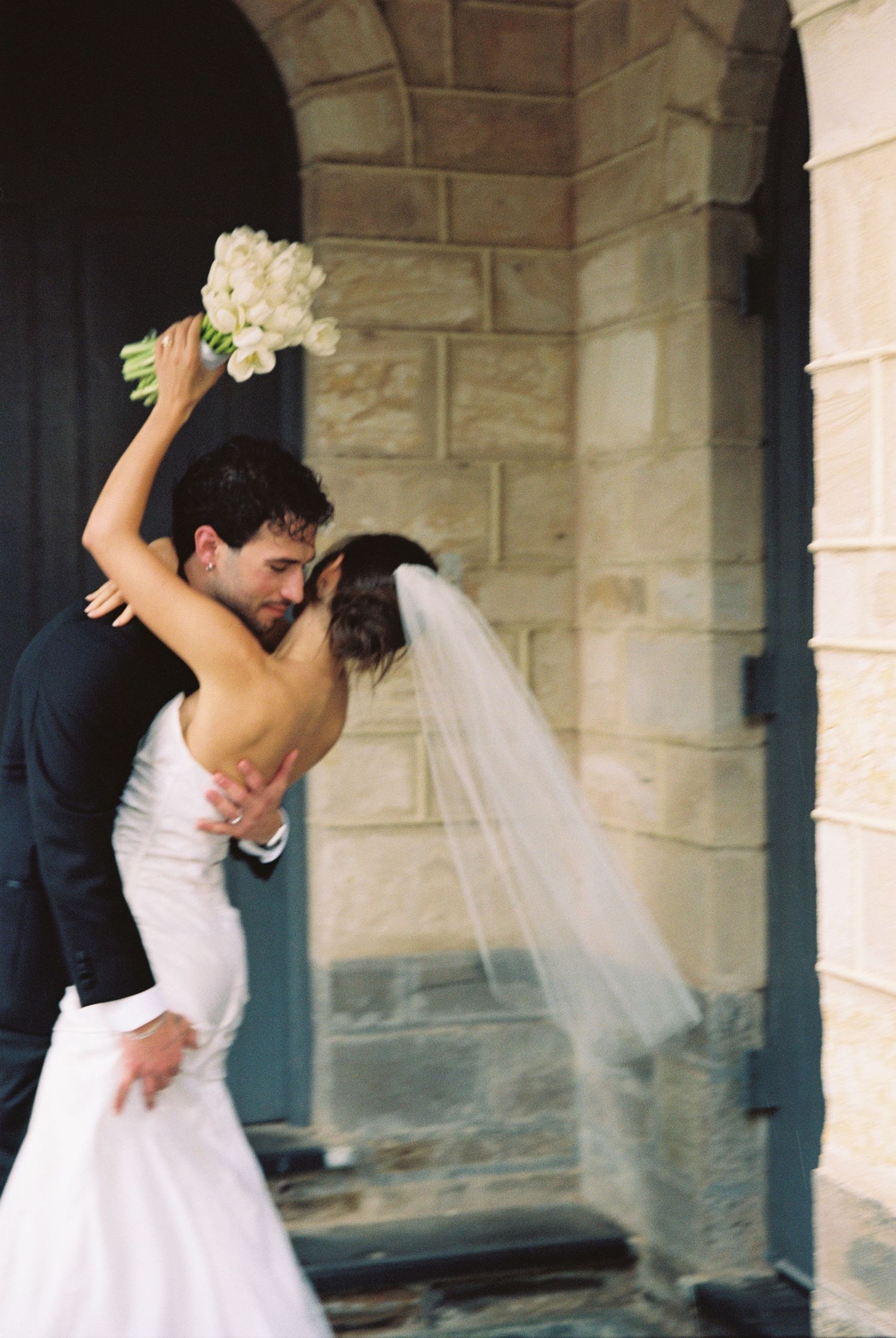 Bride and groom share a romantic dip kiss outside a stone church, bride holding white bouquet aloft.