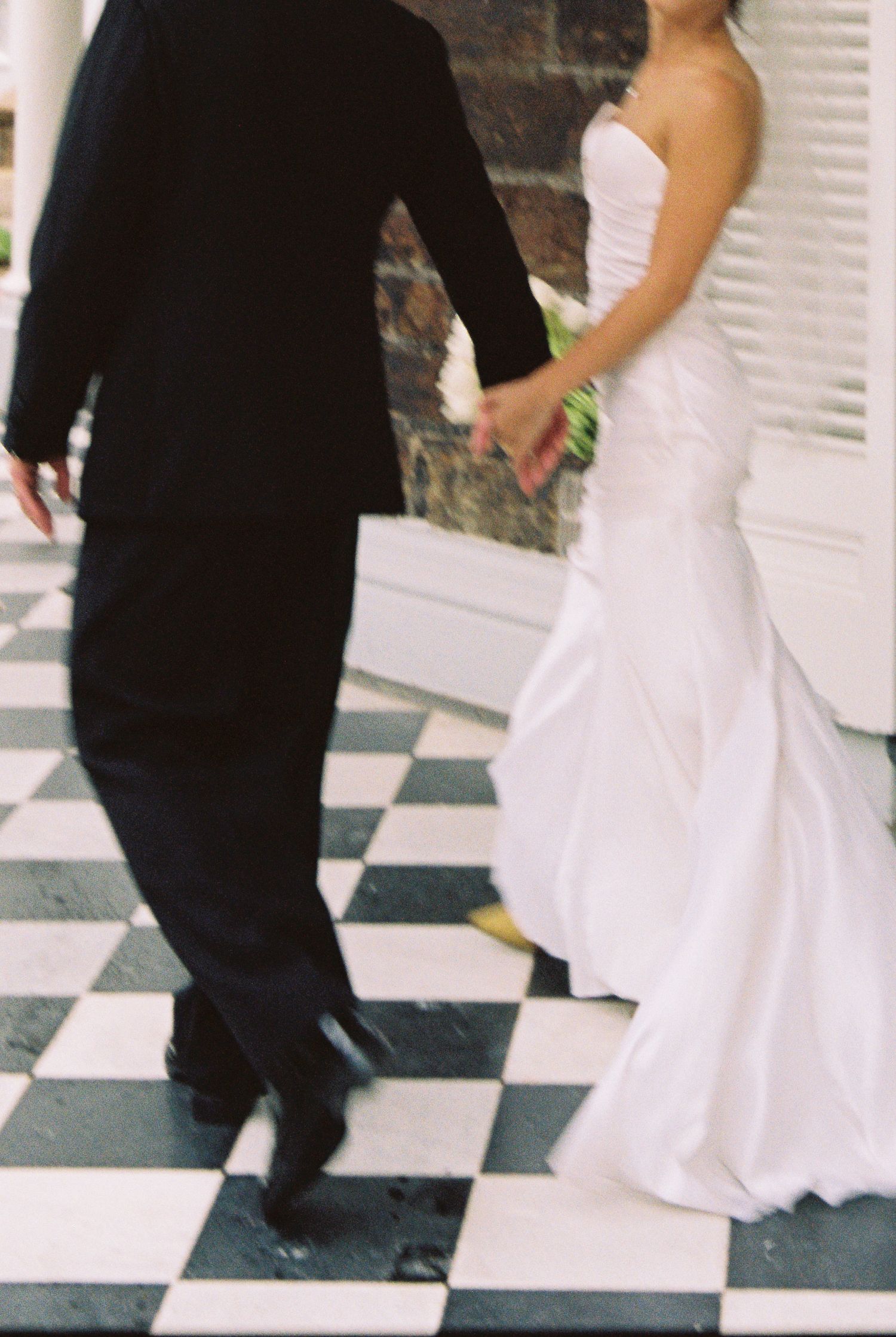 Bride in white gown and groom in black tuxedo walking hand in hand on checkered black and white floor.