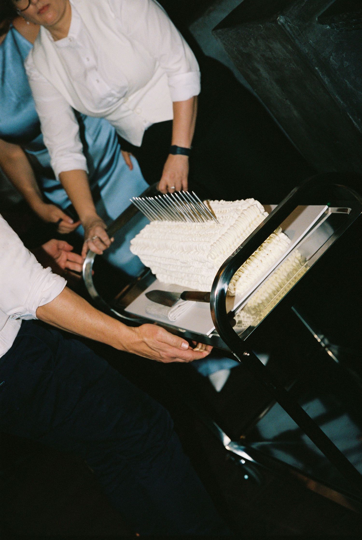 Catering staff in white shirts cutting a white frosted wedding cake on a black stand at an elegant event.