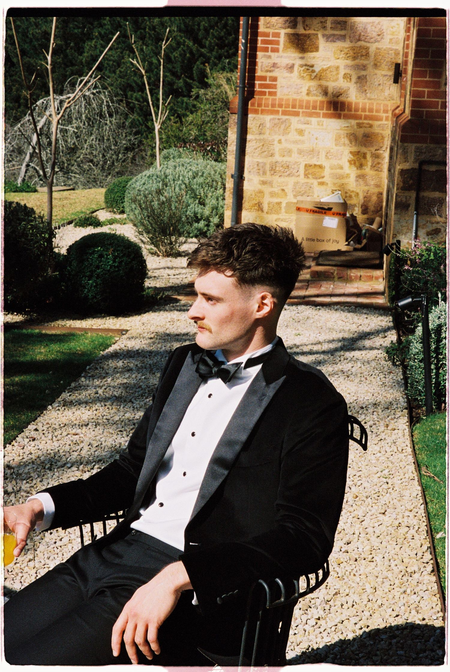 Stylish groom in black tuxedo relaxes in chair beside stone building at outdoor wedding venue.