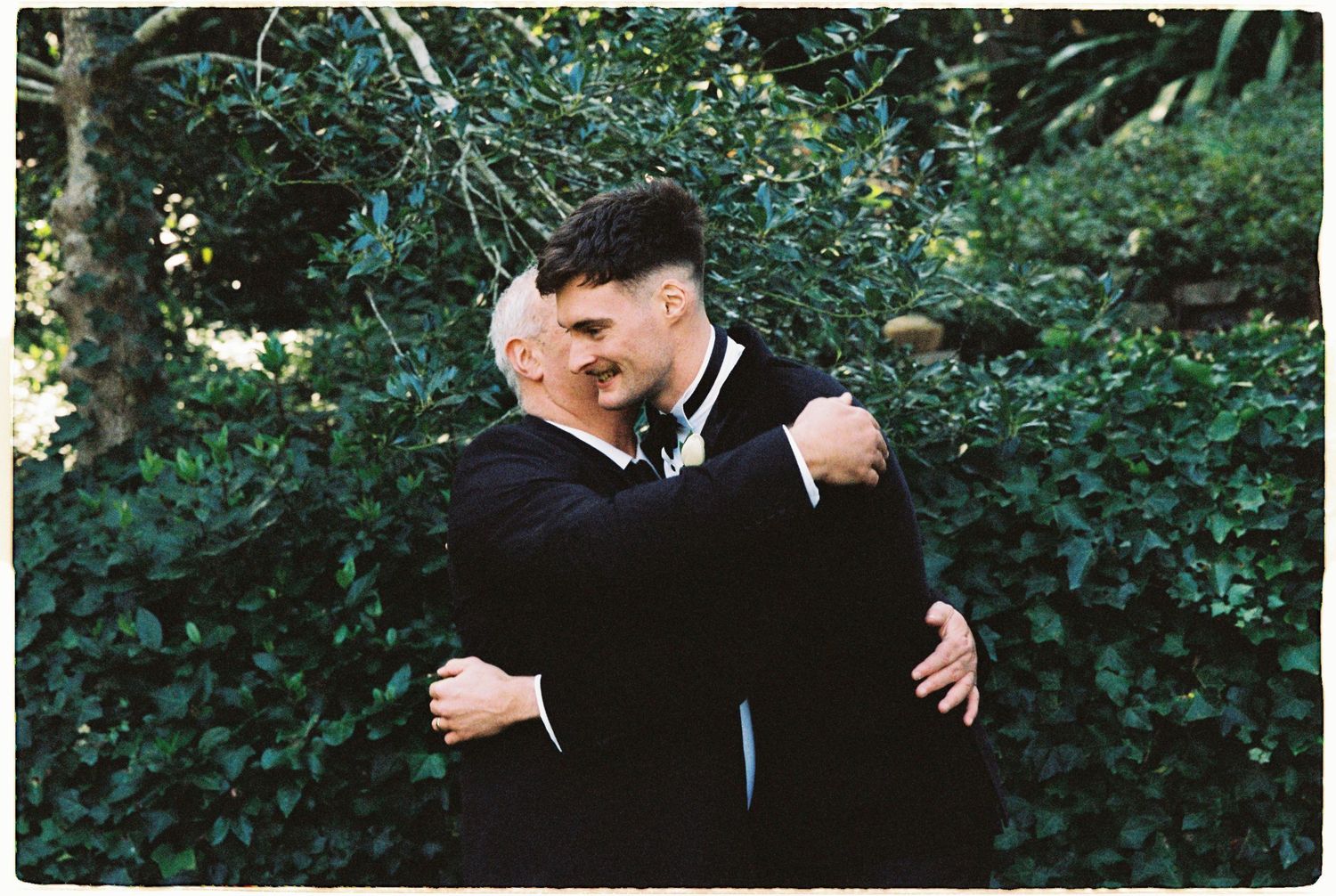 Two men in tuxedos sharing a warm embrace in front of lush greenery.