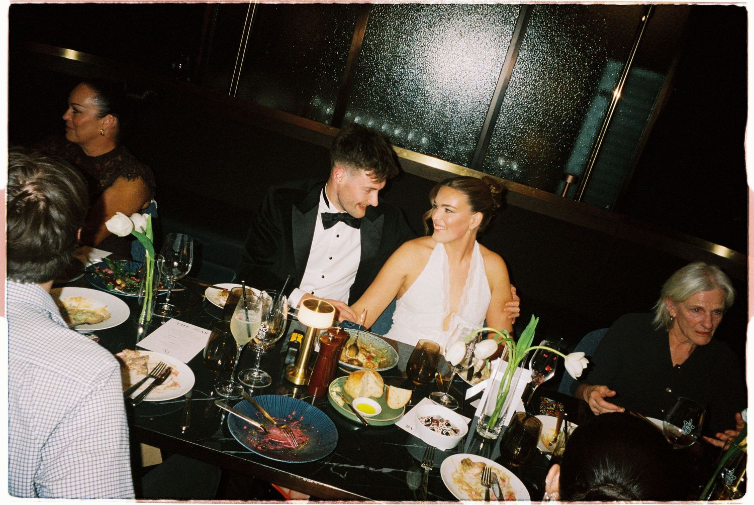 Bride and groom seated together at dark upscale wedding reception dinner table with guests nearby.
