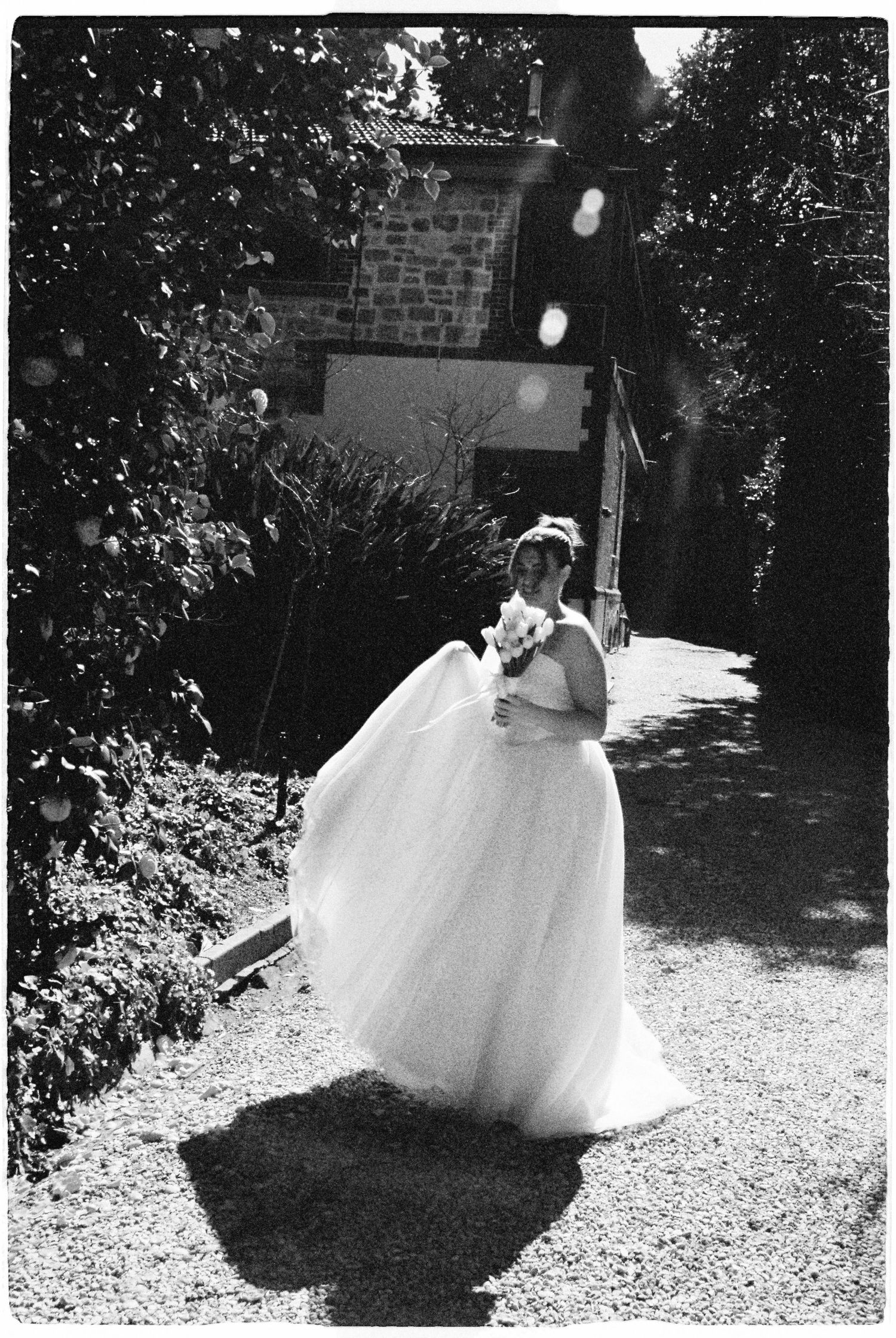 Black and white photo of a bride in a flowing white gown holding a bouquet in a sunlit garden pathway.
