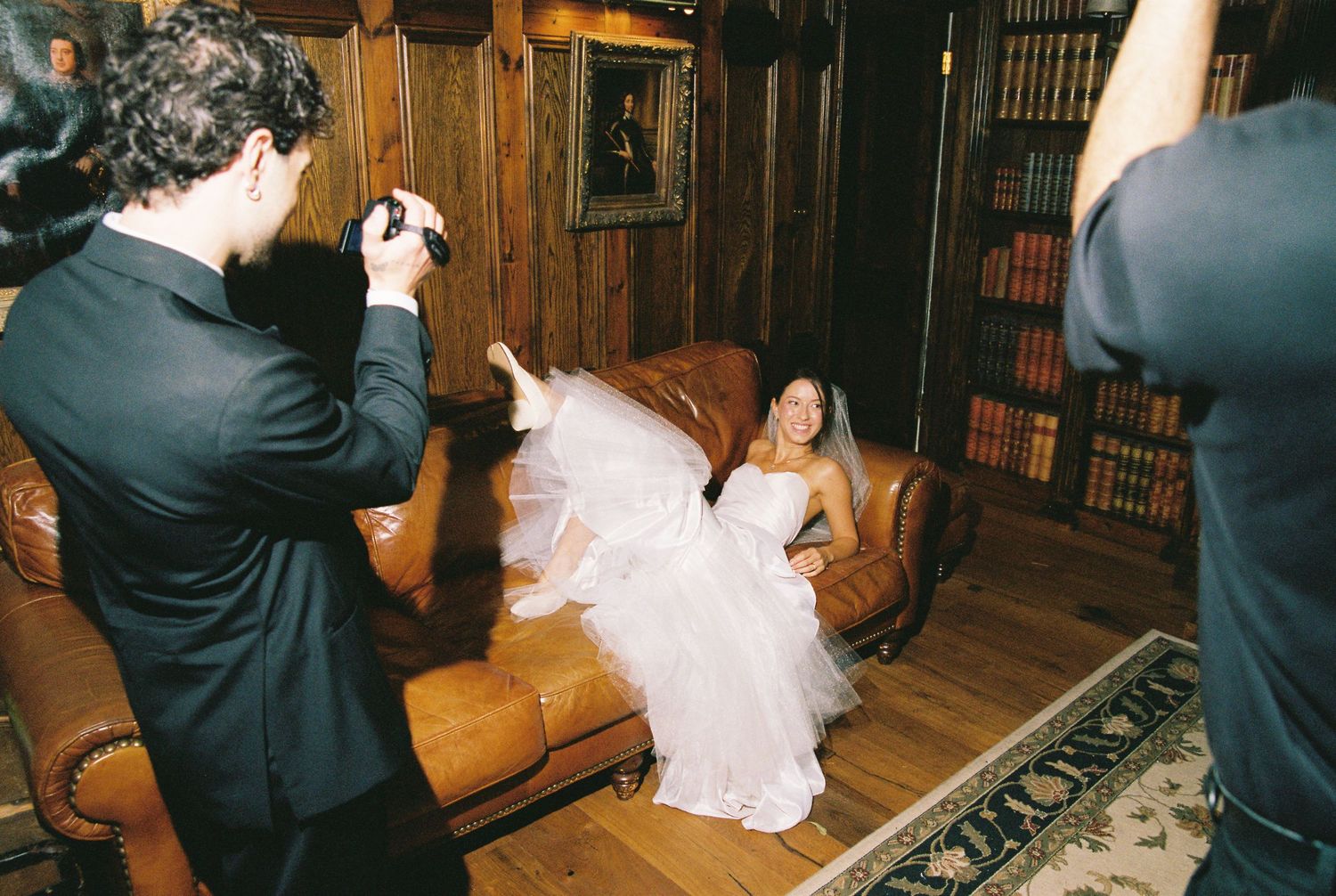 Bride in white wedding dress posing on leather sofa while photographers capture her portrait in a cozy library setting.
