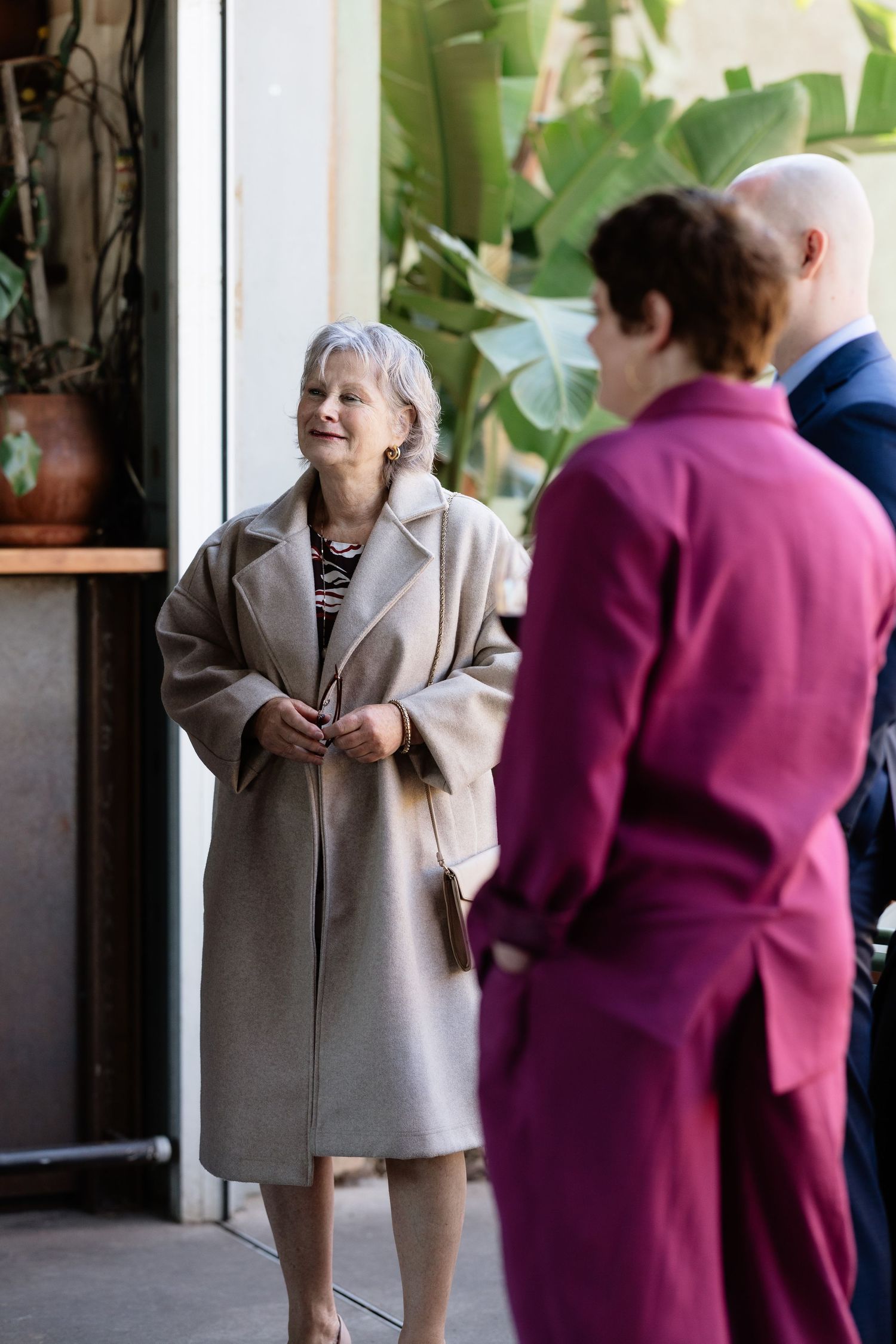 Smiling silver-haired woman in beige coat chatting with wedding guests outside a venue with lush greenery.