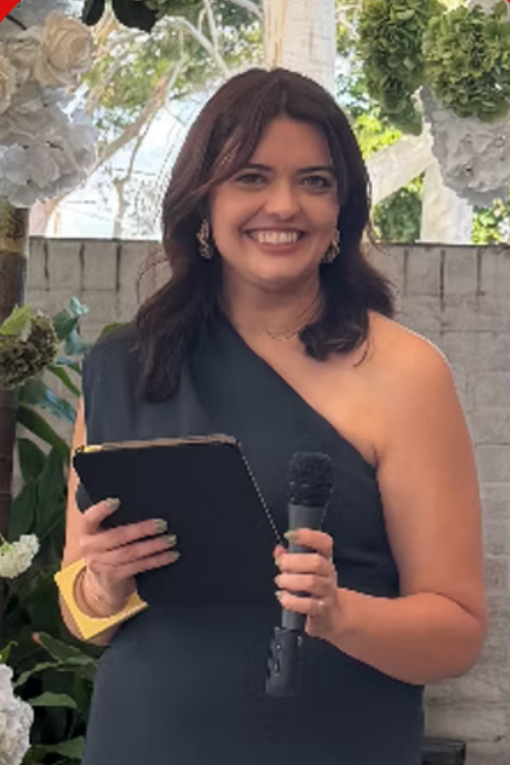 Smiling woman in gray one-shoulder dress holds microphone and book at elegant floral wedding ceremony.
