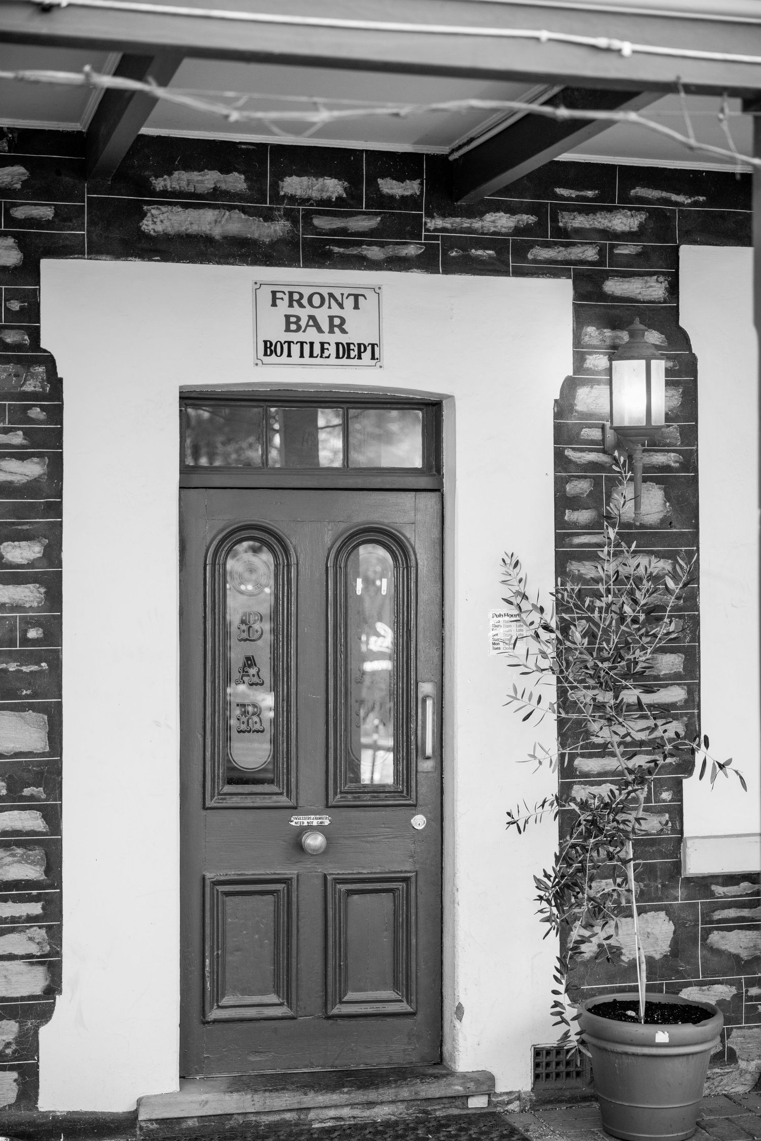 Black and white photo of a pub front bar entrance with wooden door and Bottle Dept sign, potted plant beside.