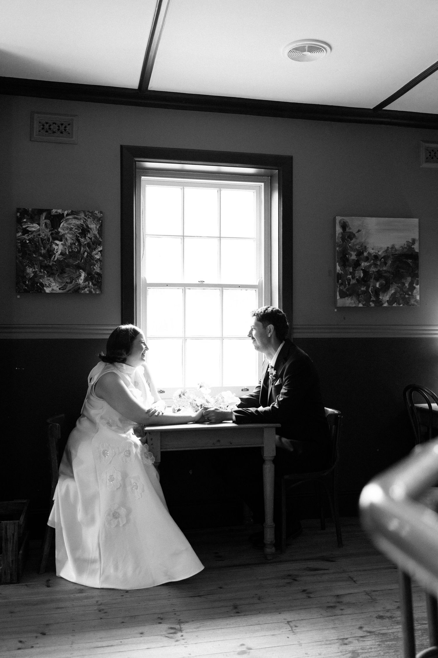 Black and white photo of a bride and groom holding hands at a table by a window in a rustic room.