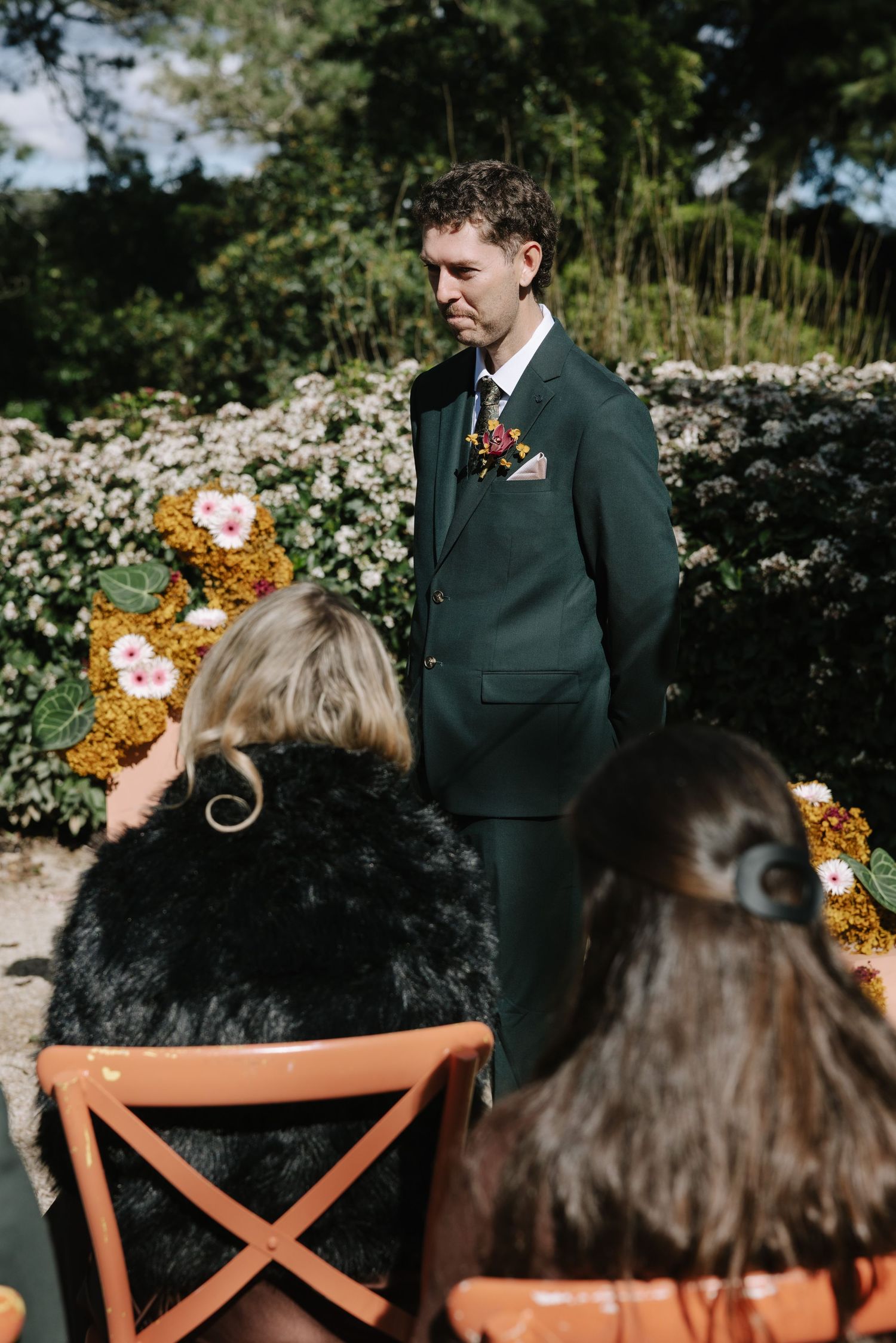 Groom in green suit with floral boutonniere stands at outdoor wedding ceremony altar surrounded by lush garden blooms.