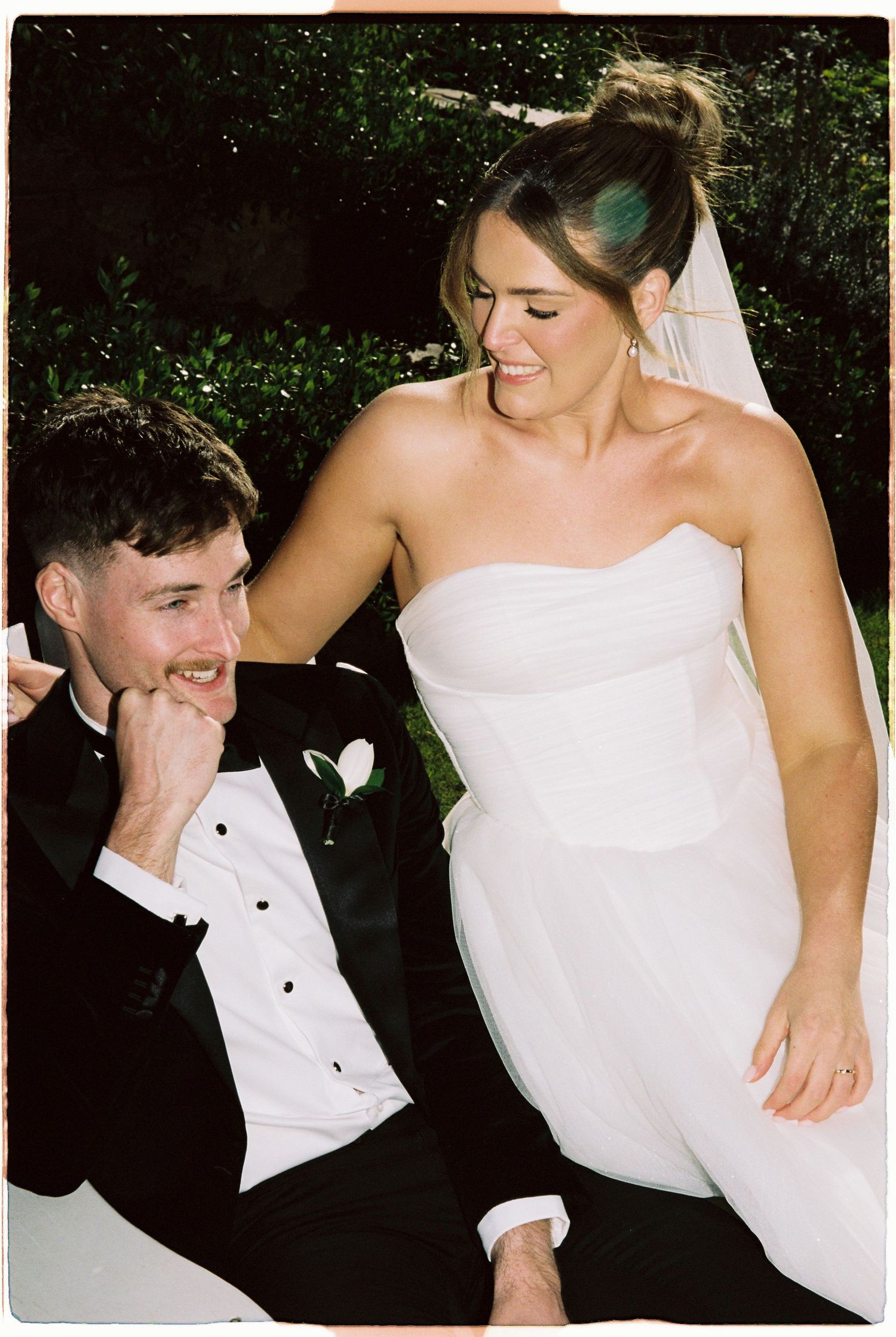 Bride in strapless white gown and groom in black tuxedo laughing together outdoors.