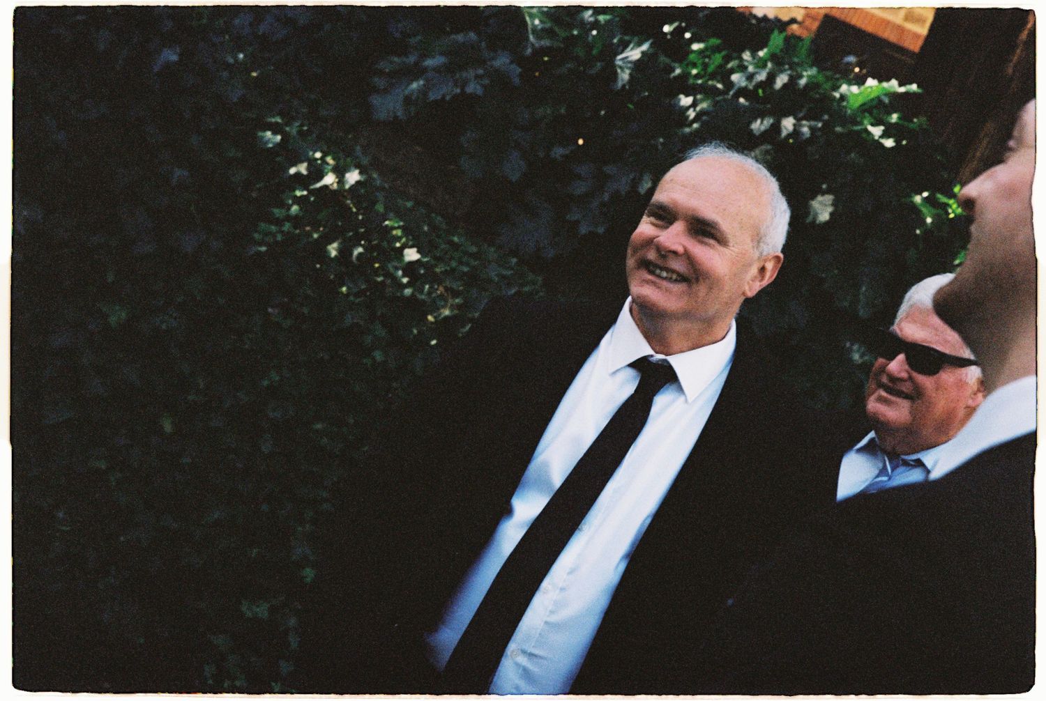 Two men in dark suits smiling and laughing together at an outdoor garden wedding event.