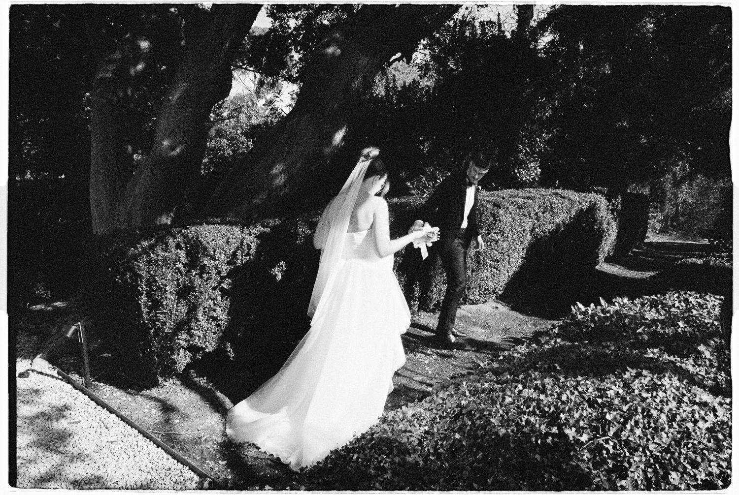 Black and white photo of a bride and groom walking through a lush garden, bride in flowing gown and veil.